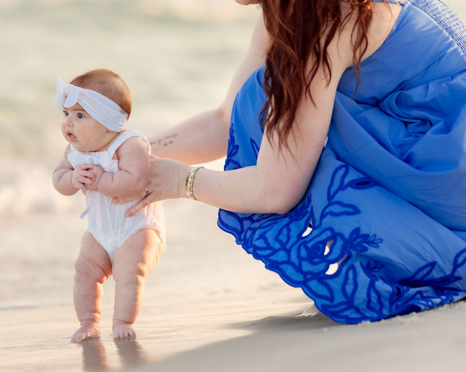 Baby girl in white outfit with headband takes first steps on beach, guided by mother in blue floral dress.