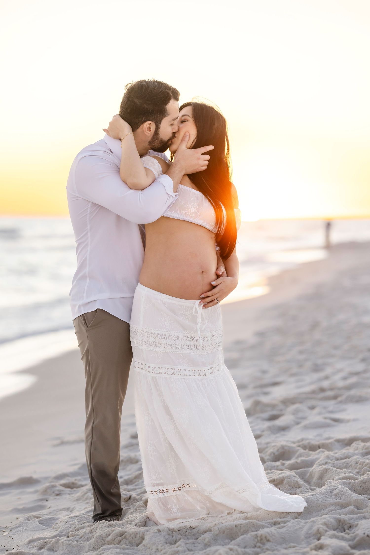 A romantic couple embraces on a sandy beach at sunset with soft golden light illuminating their tender moment.