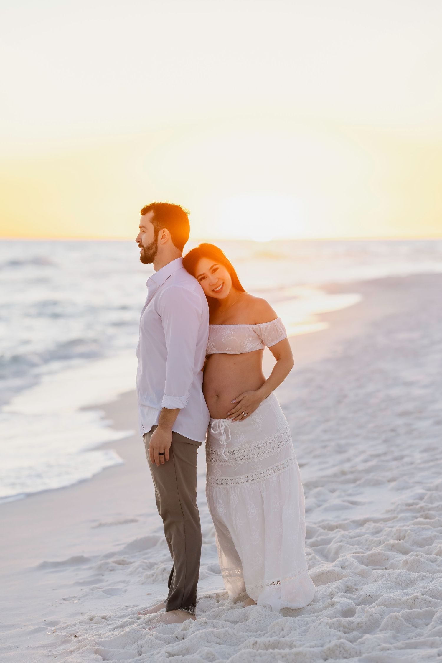 An expectant couple poses together on a white sandy beach during golden hour.