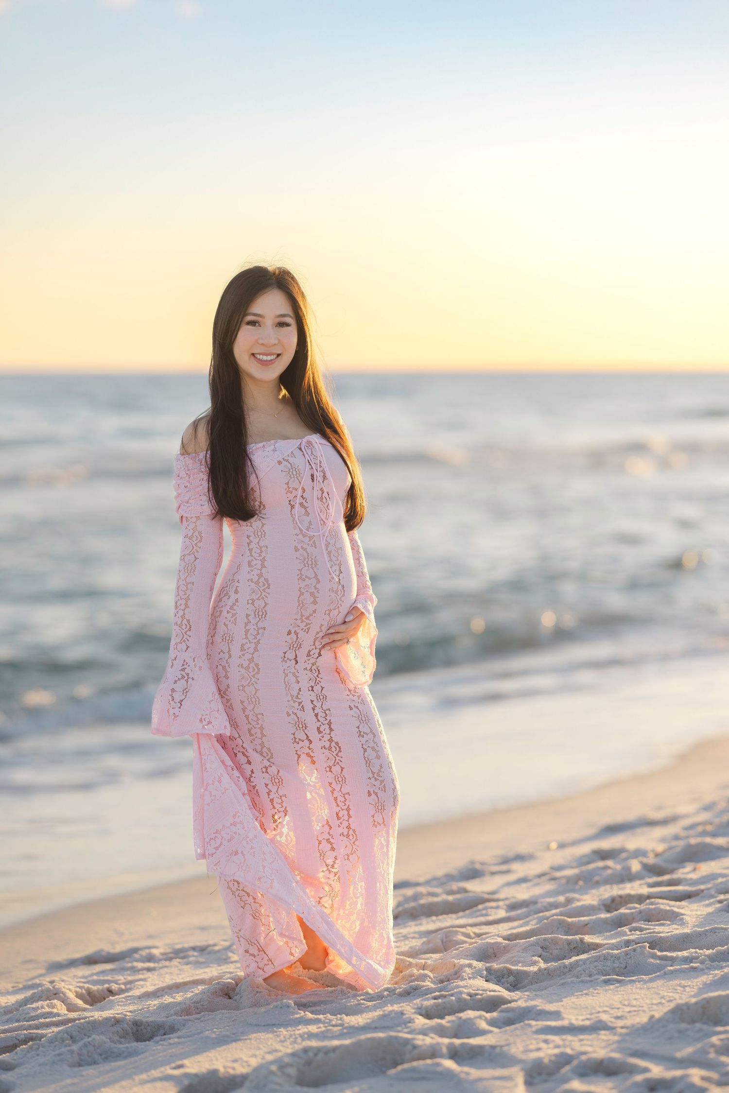 A pink lace dress flows in the breeze as someone walks along a sandy beach at sunset by the ocean.