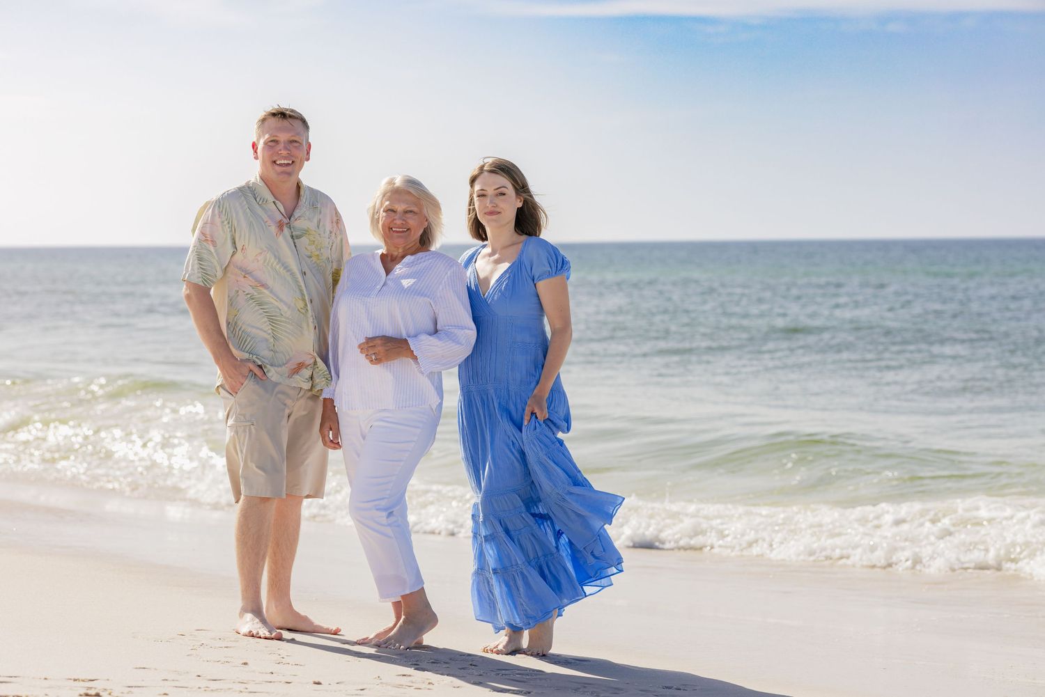 Three people smiling together on a sunny beach with ocean waves in the background.