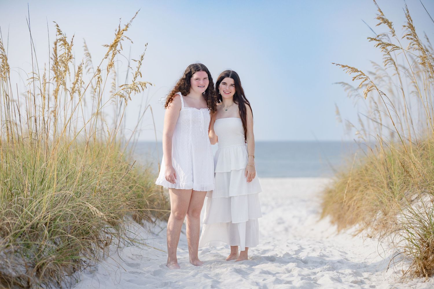Two smiling young women in white outfits pose together on a sandy beach path with sea oats behind them.