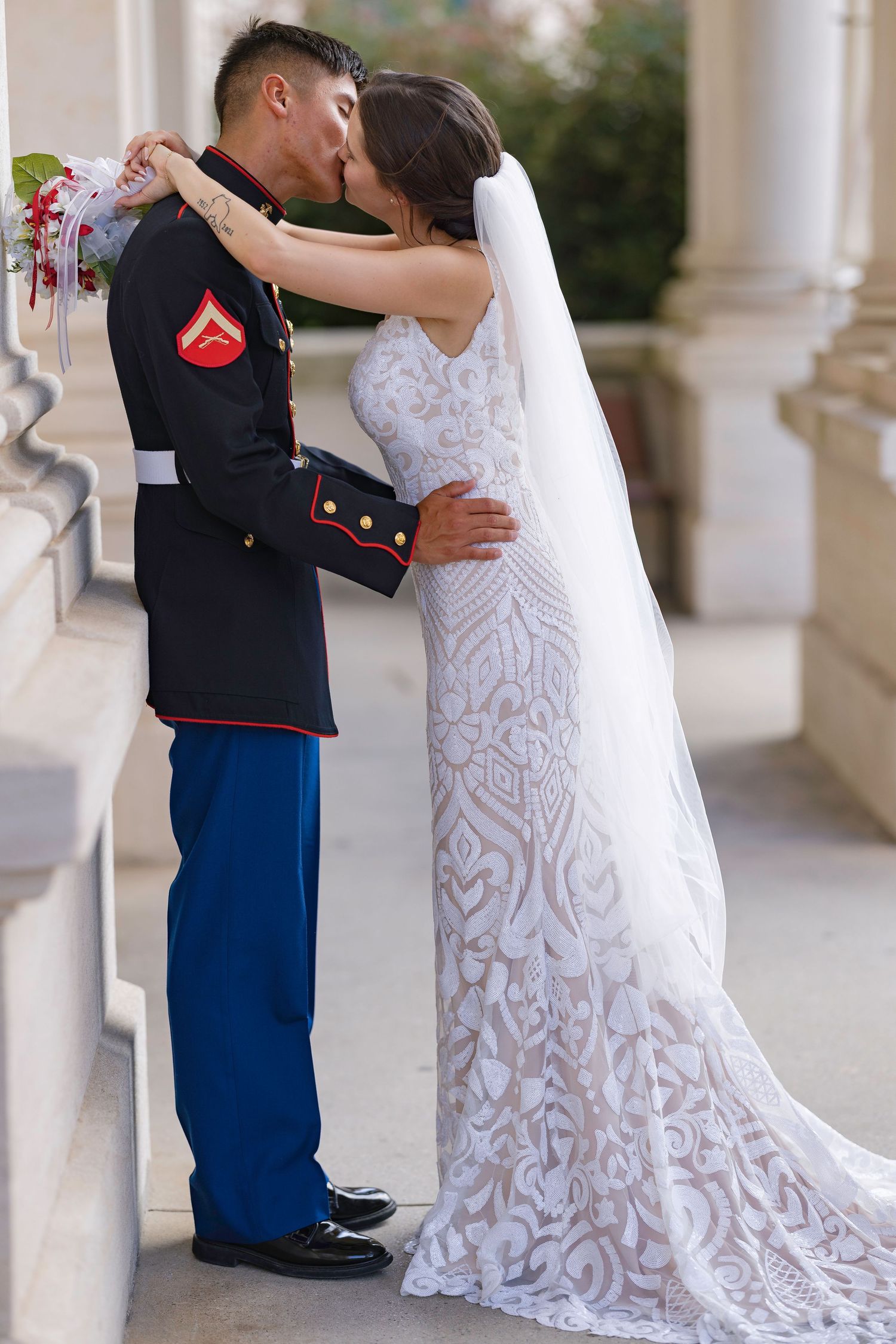 Marine in dress uniform kisses bride in lace wedding gown between columns at elegant outdoor ceremony.