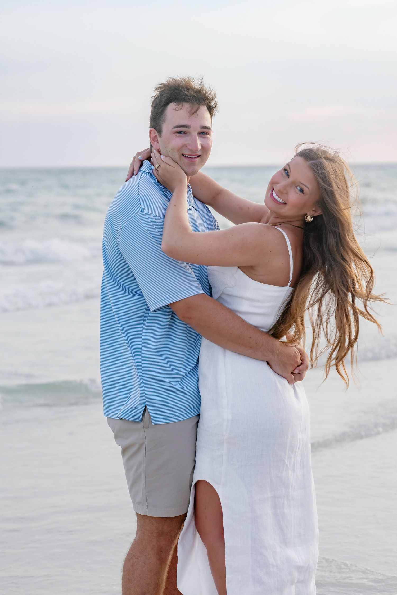 Happy couple embracing on beach at sunset, man in blue shirt and woman in white dress.