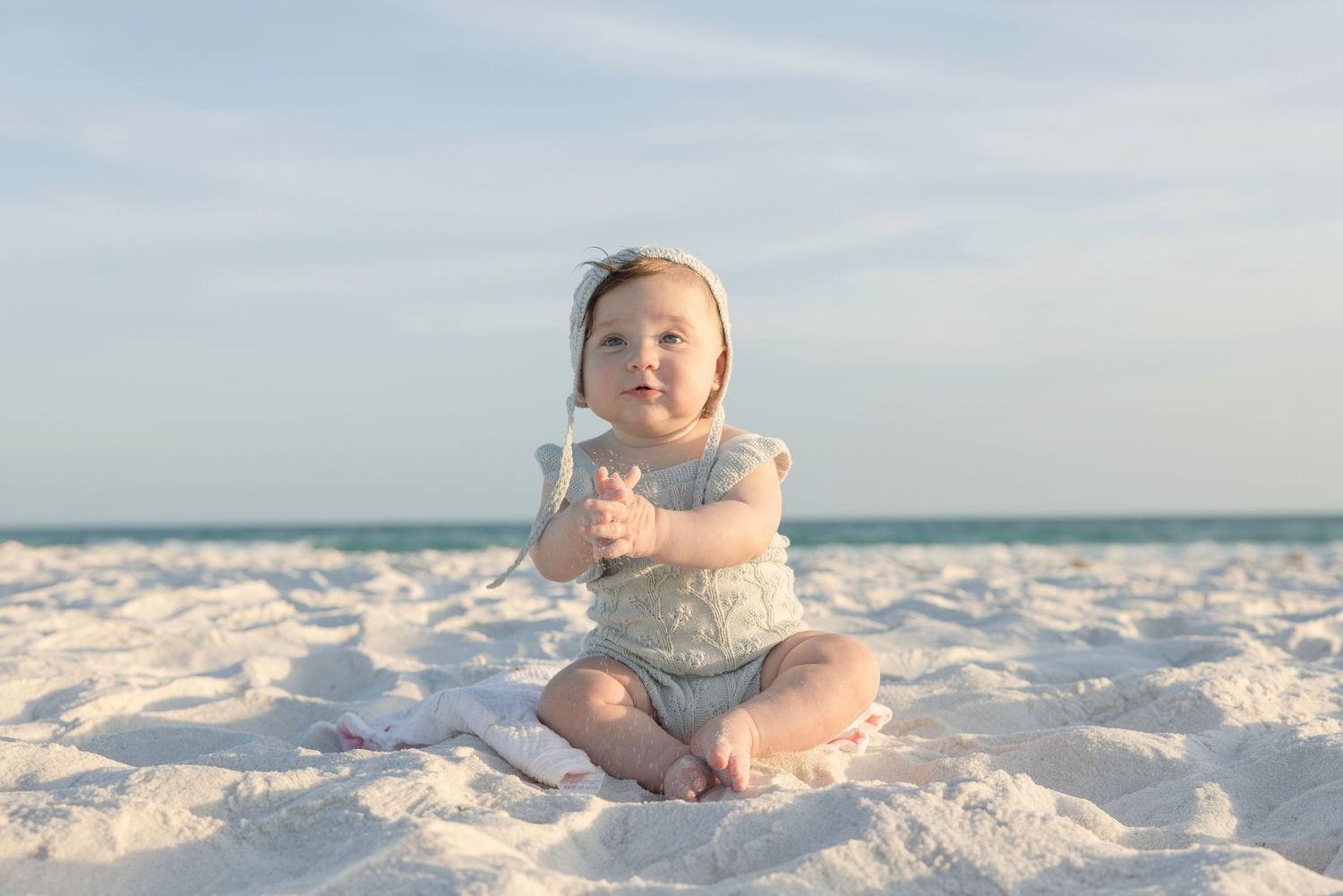 Smiling baby girl sitting on white sand beach in gray romper, with ocean and blue sky in the background.