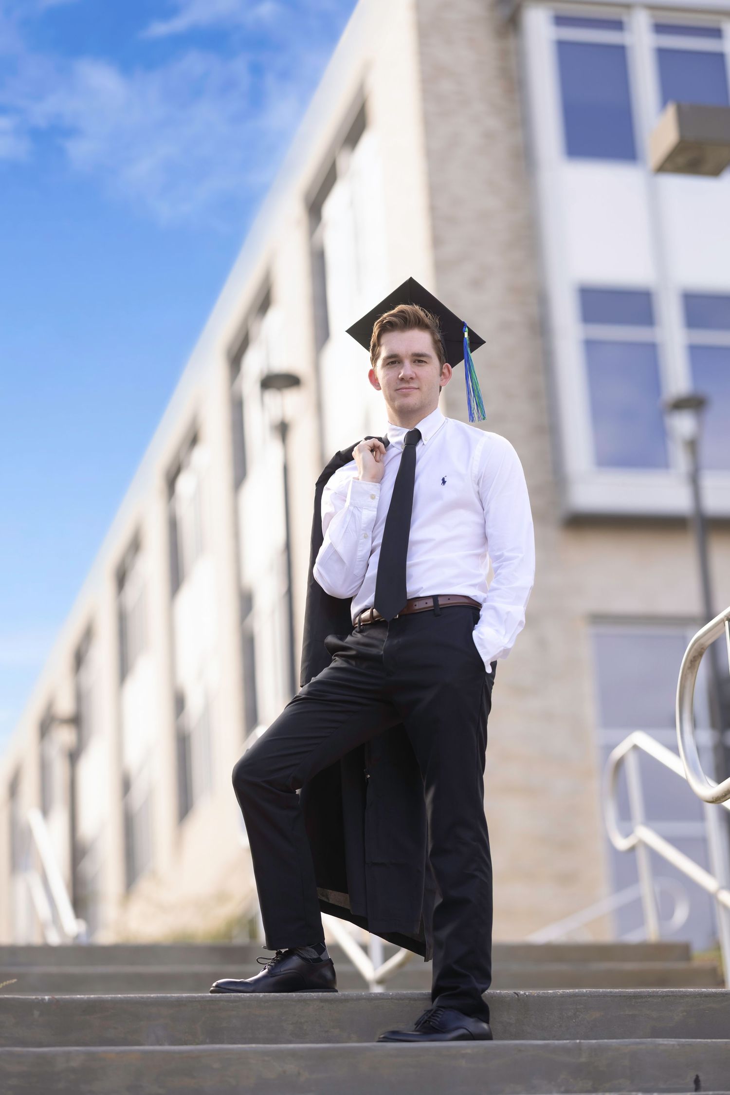 Young man in white shirt, tie, and graduation cap poses confidently on steps outside a modern building.