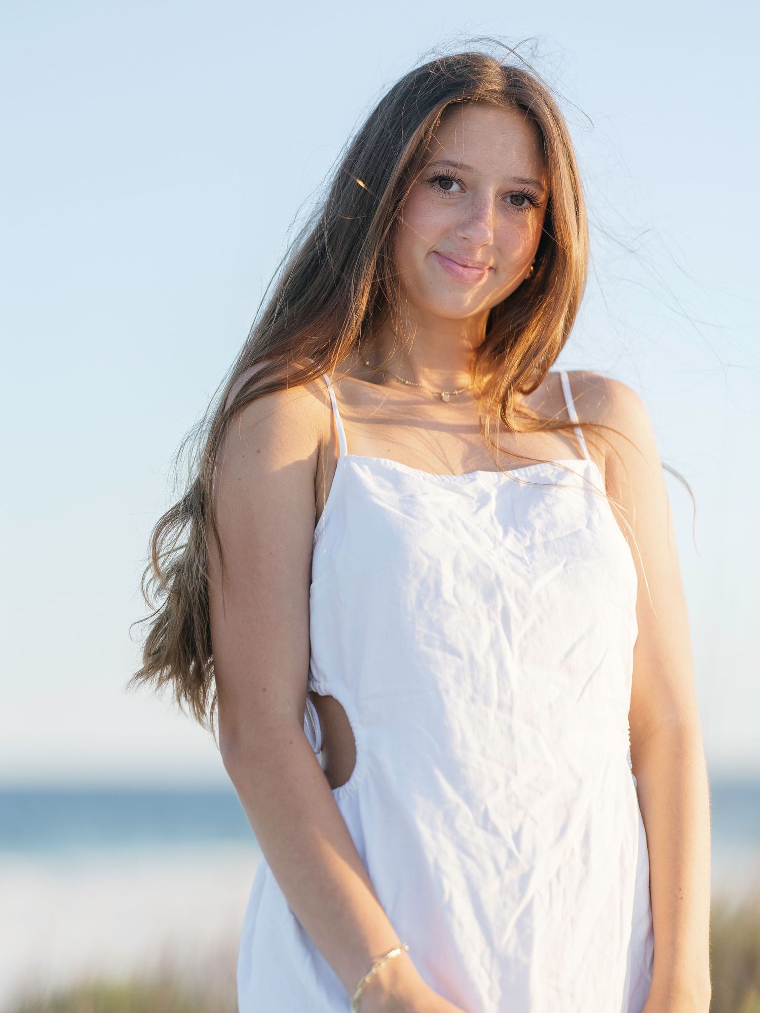 Woman in white sundress poses against bright sky with windswept hair.