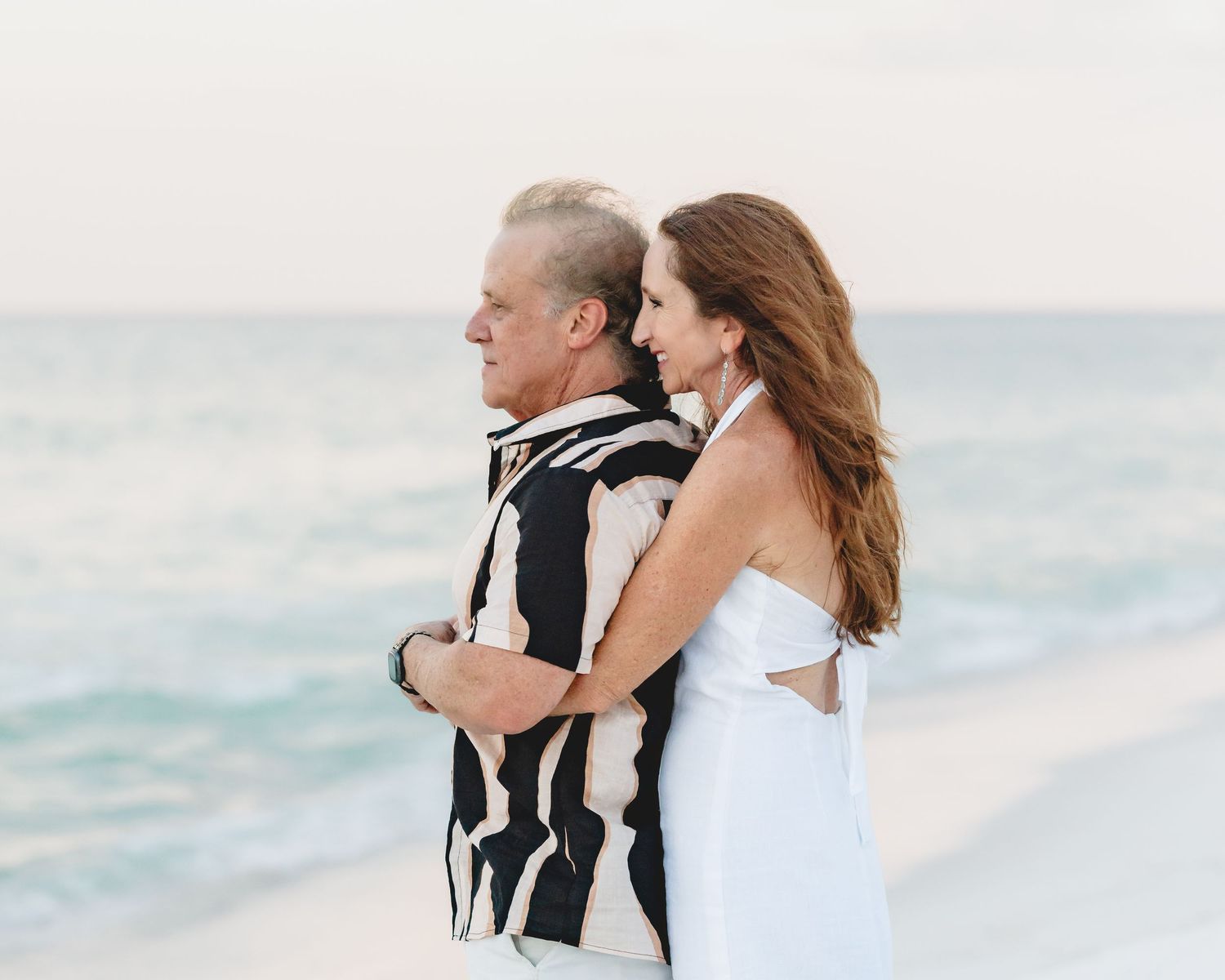 Couple embraces romantically on beach, man in black and white shirt with woman in white dress at sunset.