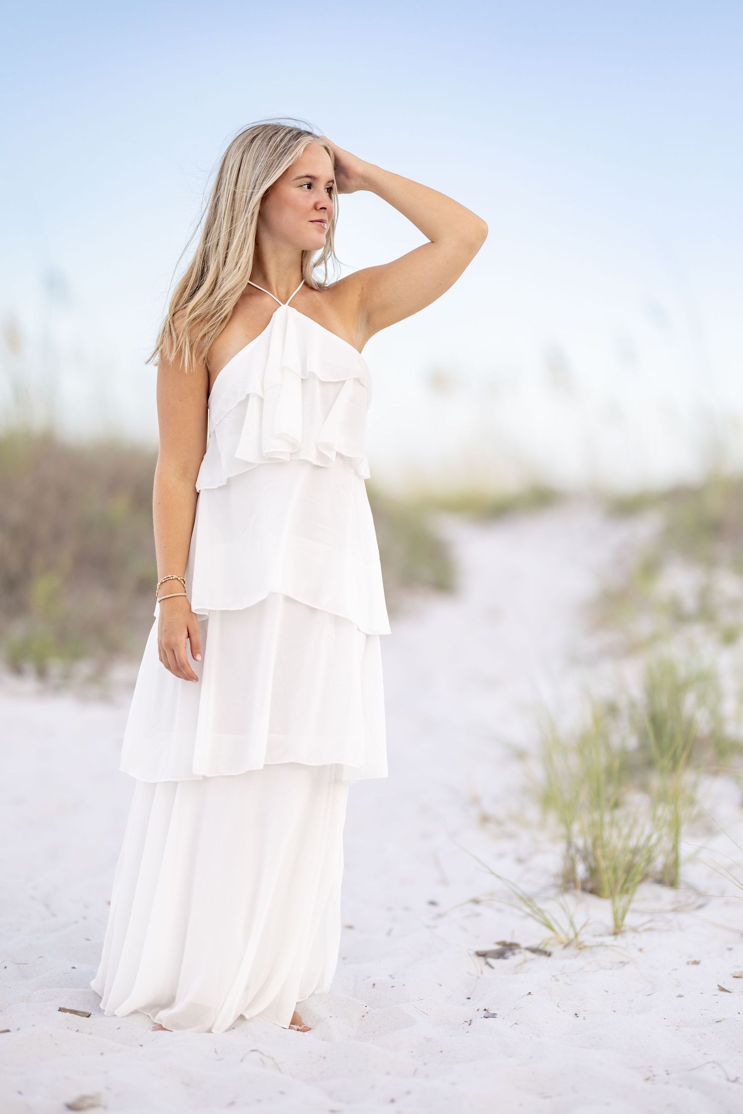 Young woman in white tiered maxi dress standing on sandy beach, hand raised to head, looking away.