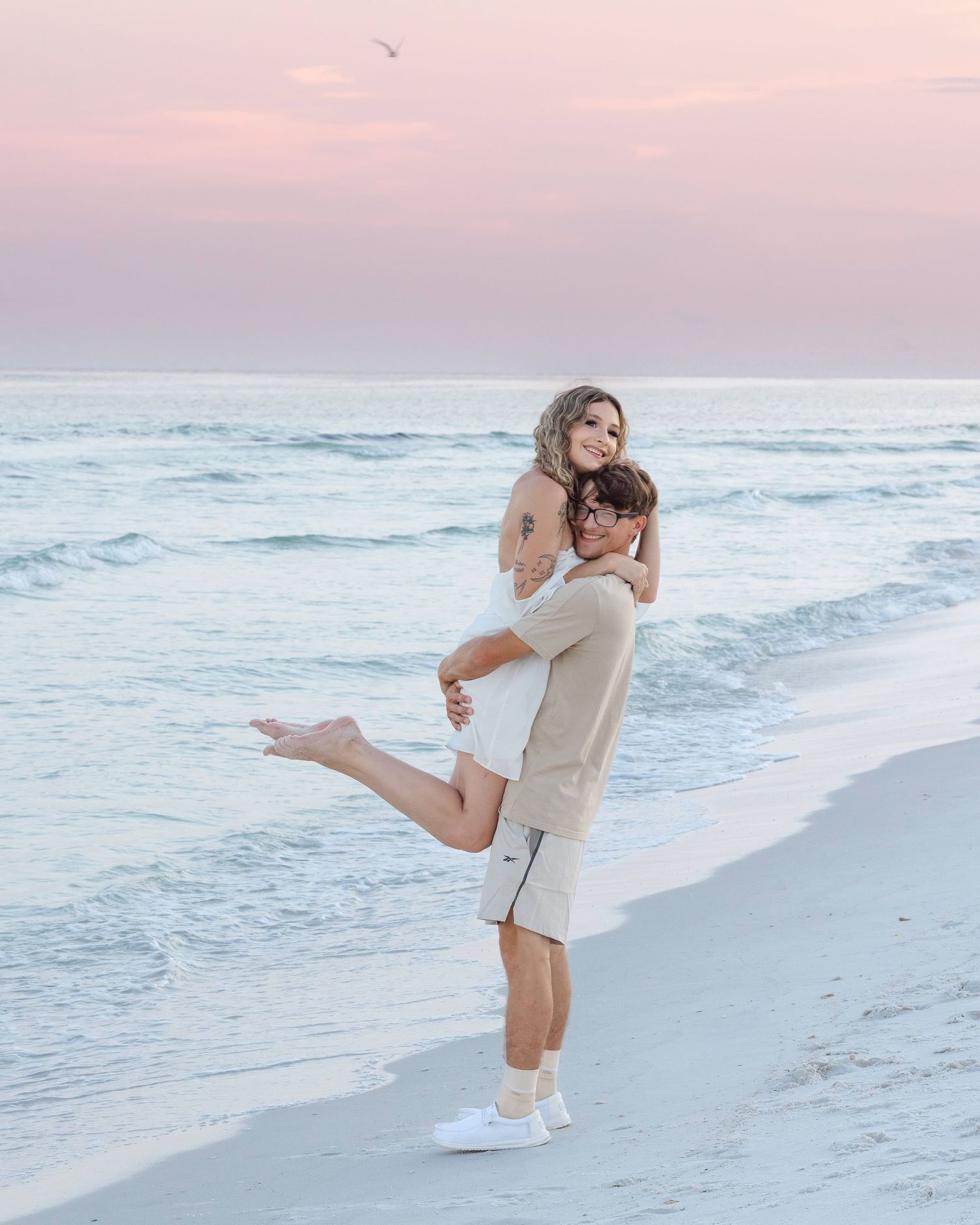 A playful couple in light colored outfits dances together on a beach at sunset.
