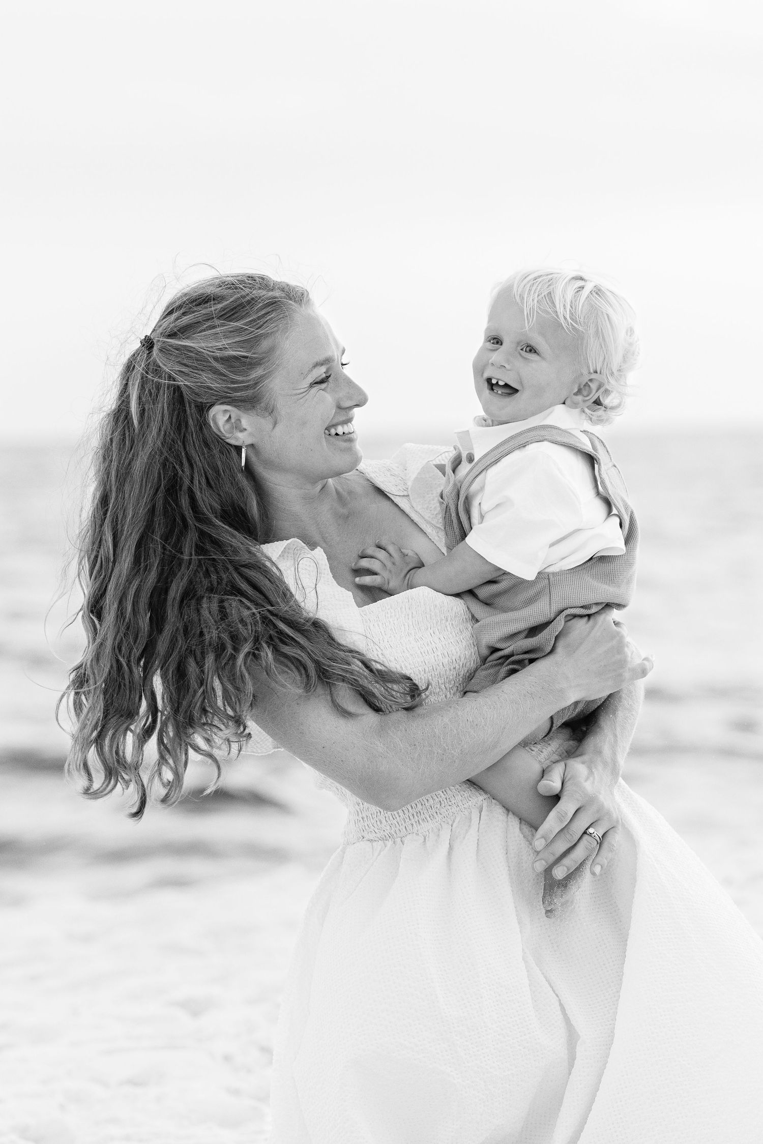 Black and white photo of parent and young child laughing together joyfully on a sunny beach.
