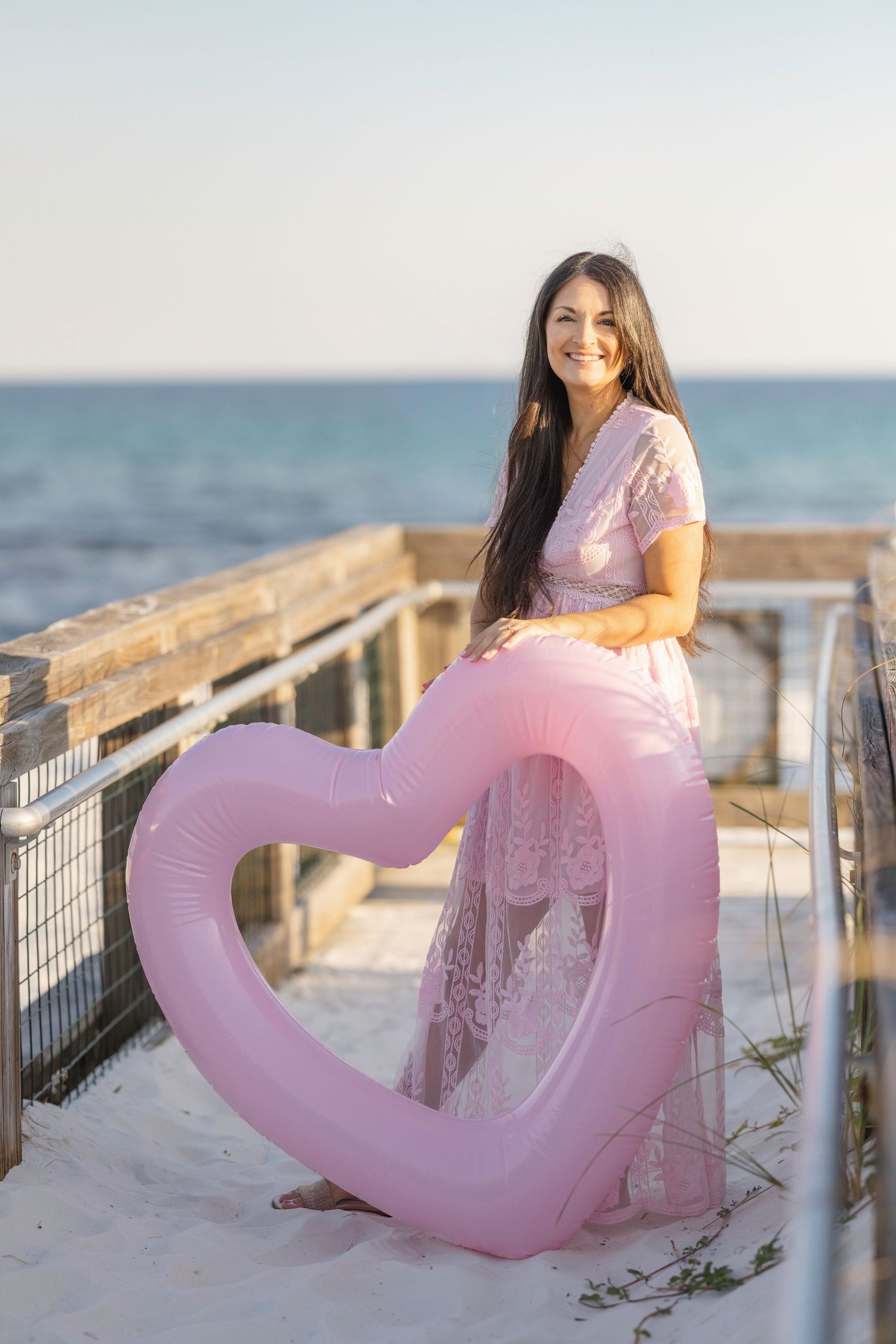 Pink inflatable heart serves as a prop for a beachside photography session on a boardwalk at sunset.