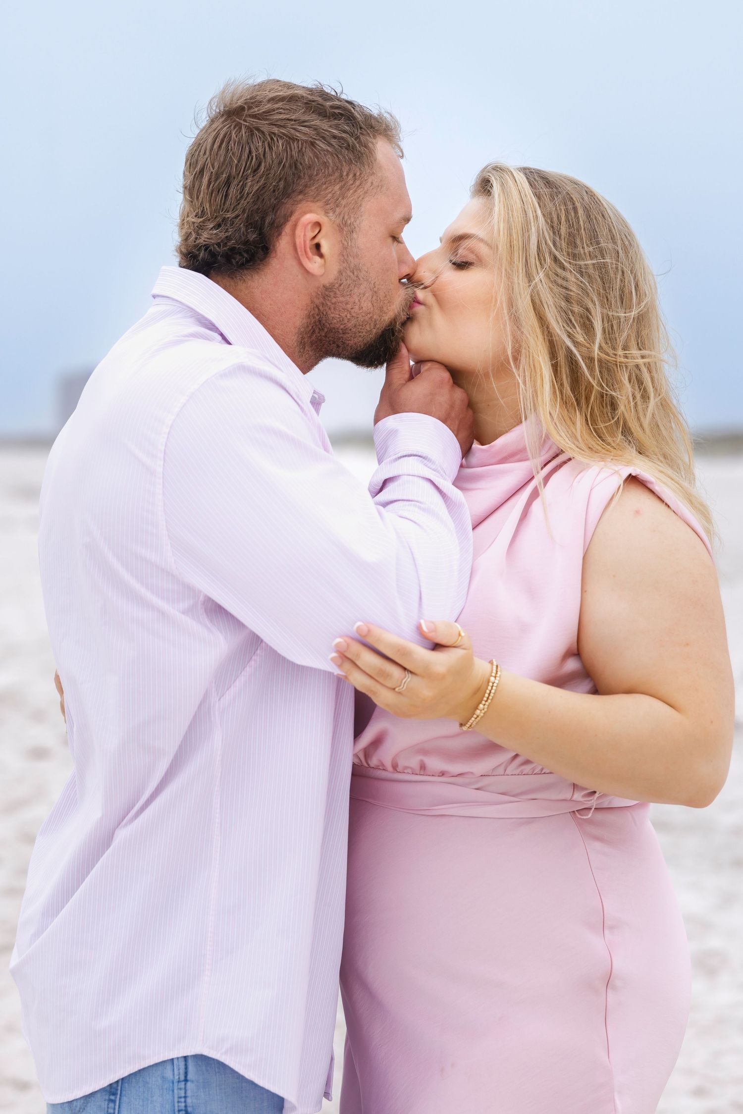 Romantic couple sharing a kiss on the beach, man in white shirt and woman in pink dress.