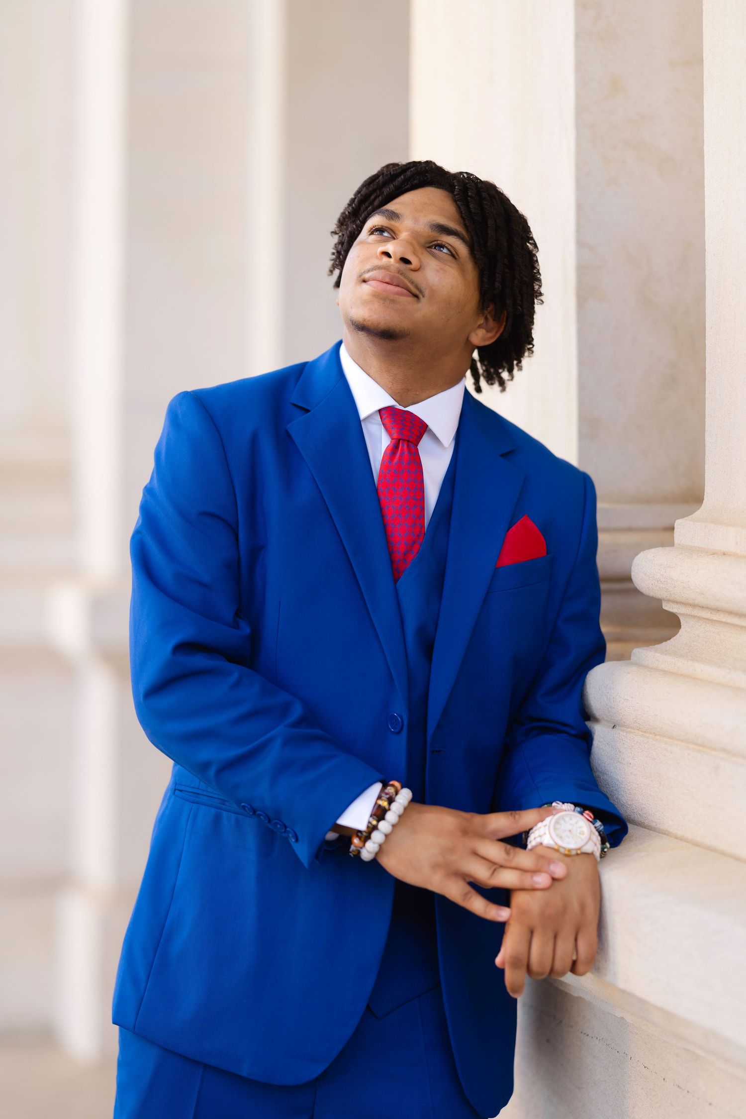 Young man in blue suit with red tie and pocket square leaning against white columns, looking upward confidently.