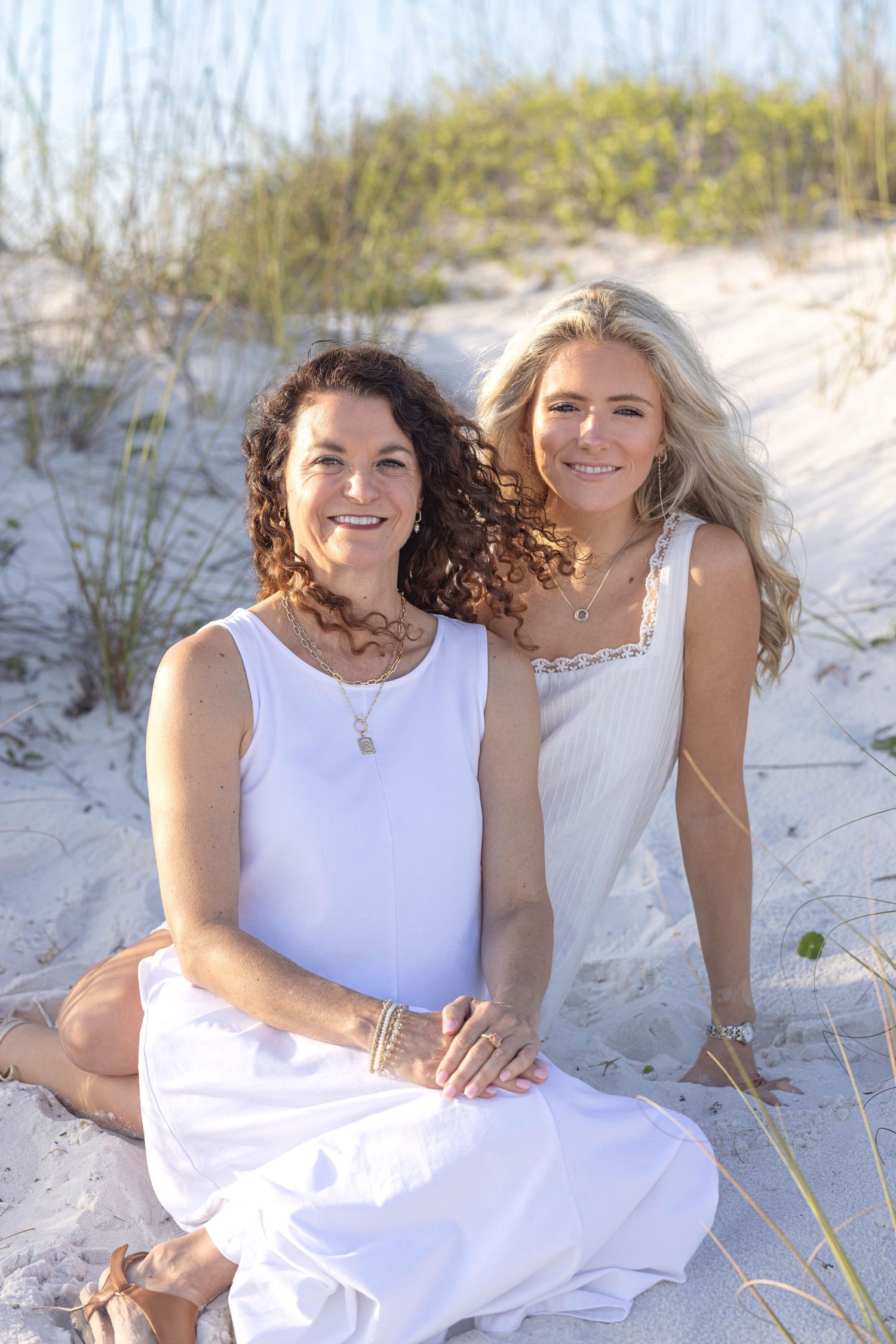 Two women in white dresses sit together on sandy beach dunes, smiling warmly in golden sunlight.