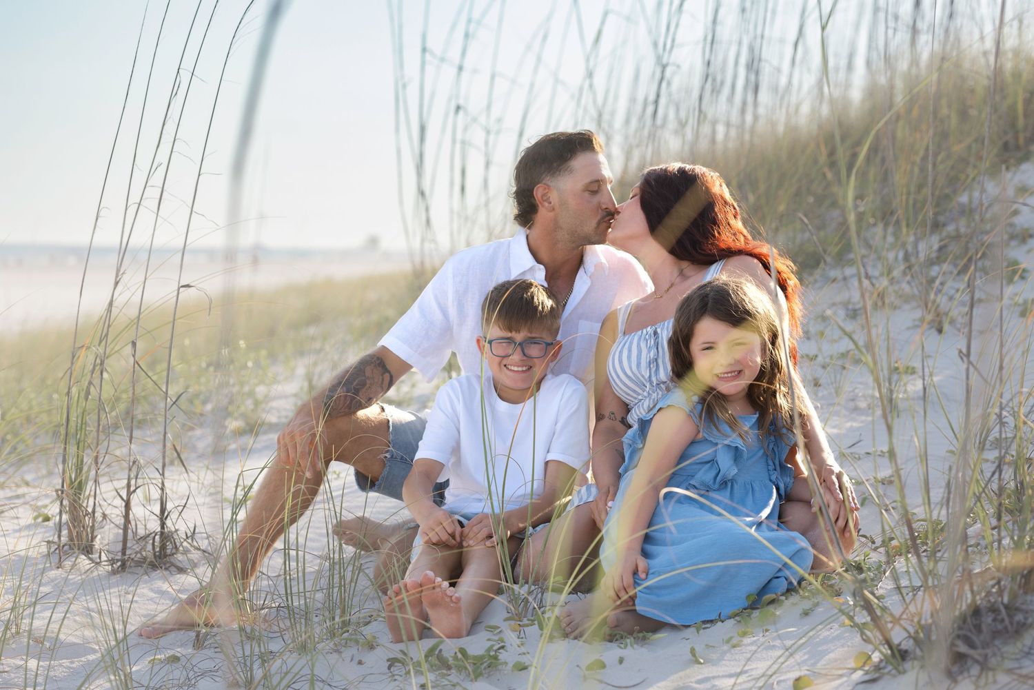 Family of four sitting on beach sand dunes among sea grass during golden hour.