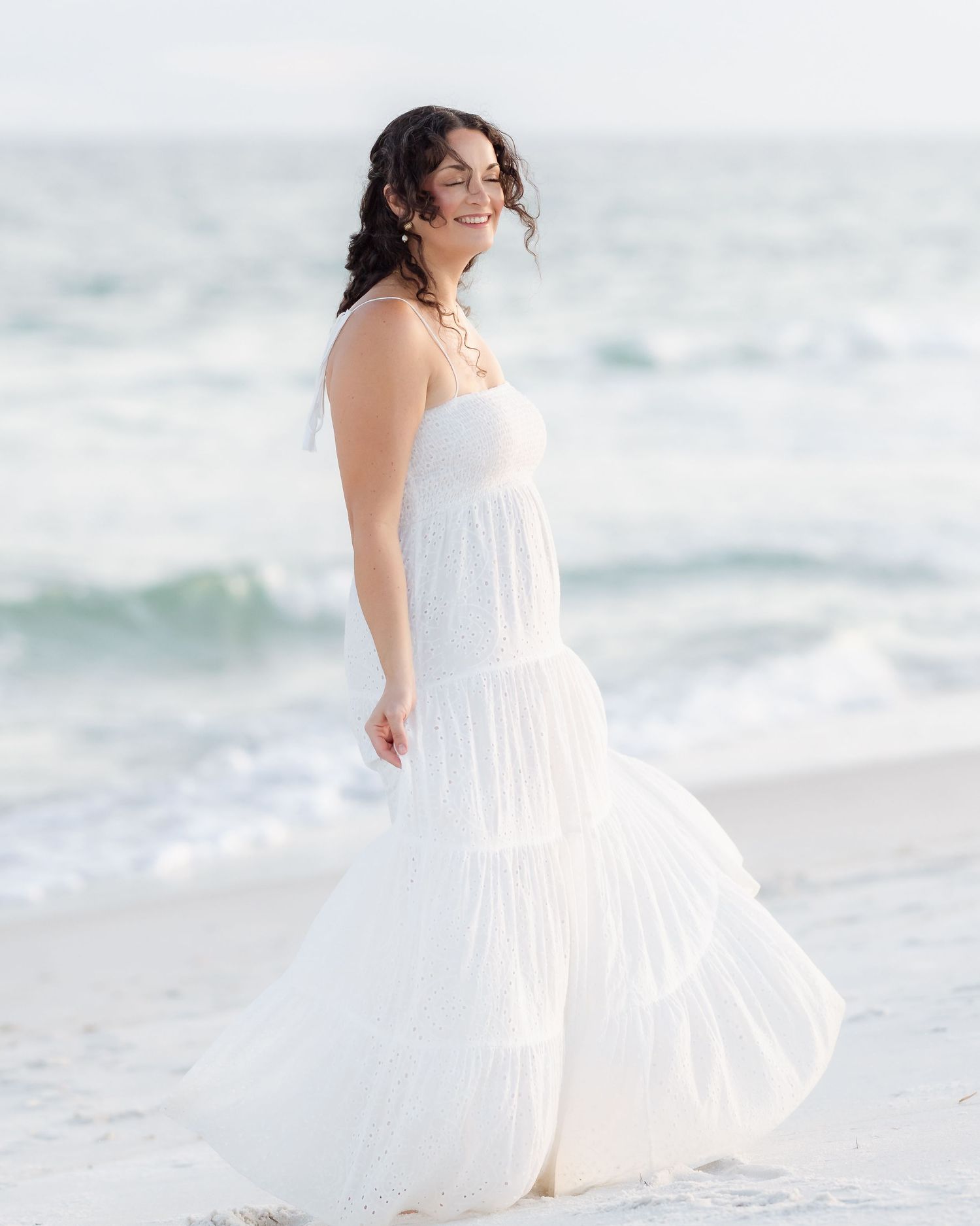 Joyful woman in flowing white tiered dress posing on beach with ocean waves behind.