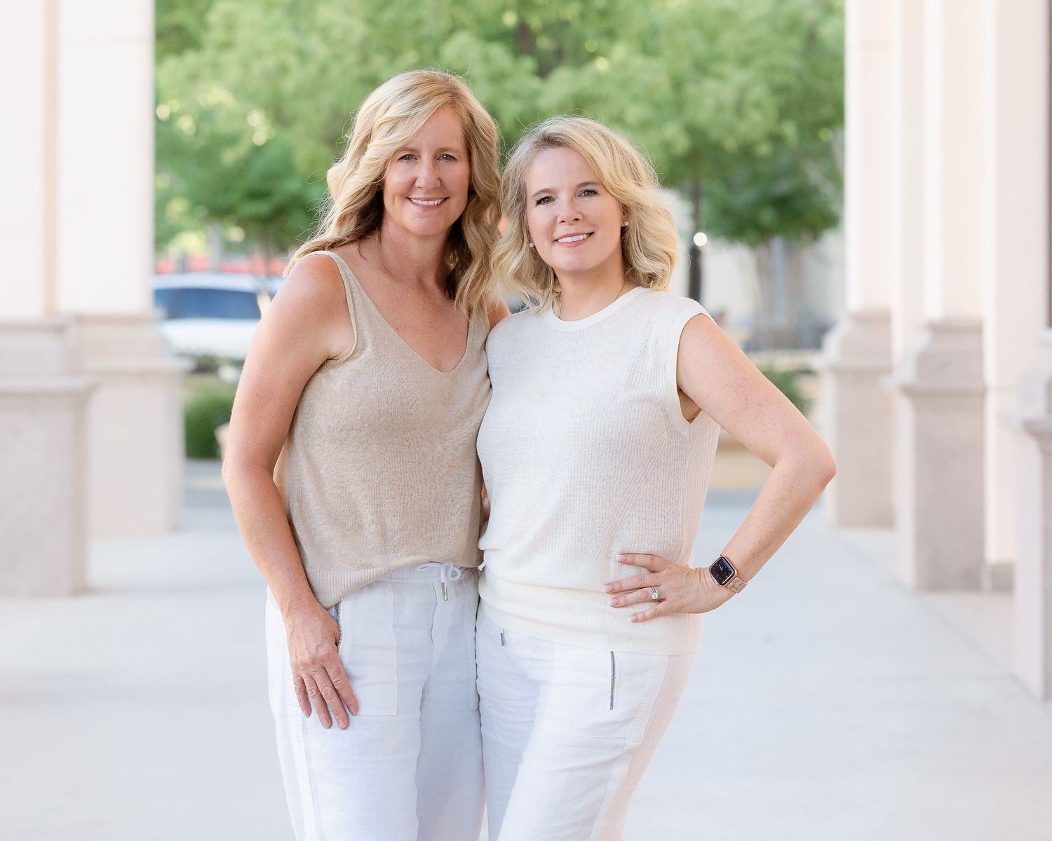 Two smiling blonde women in casual white and beige outfits pose together outdoors near elegant columns.