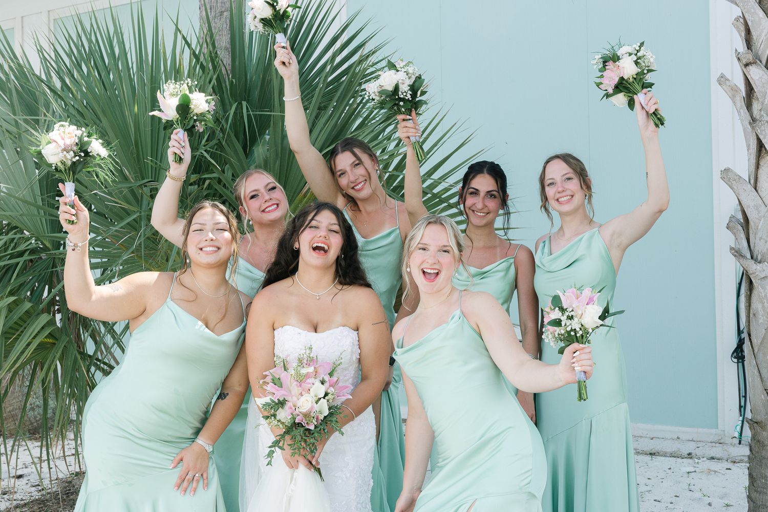 Bridesmaids in mint green dresses pose joyfully with floral bouquets on beach.