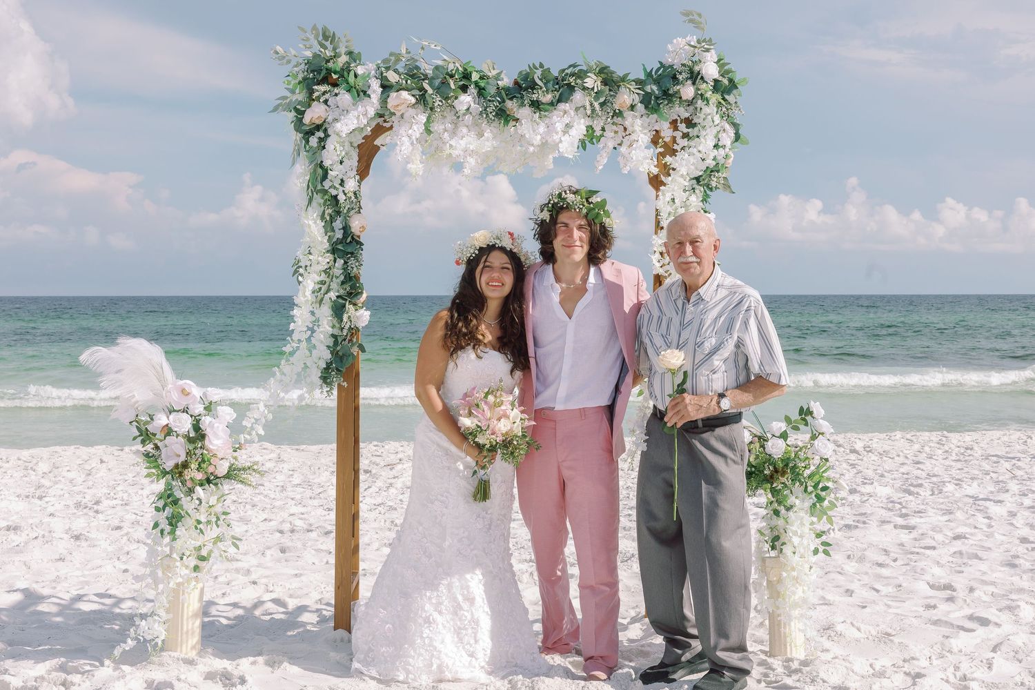 Three people pose under floral arch at beach wedding ceremony.