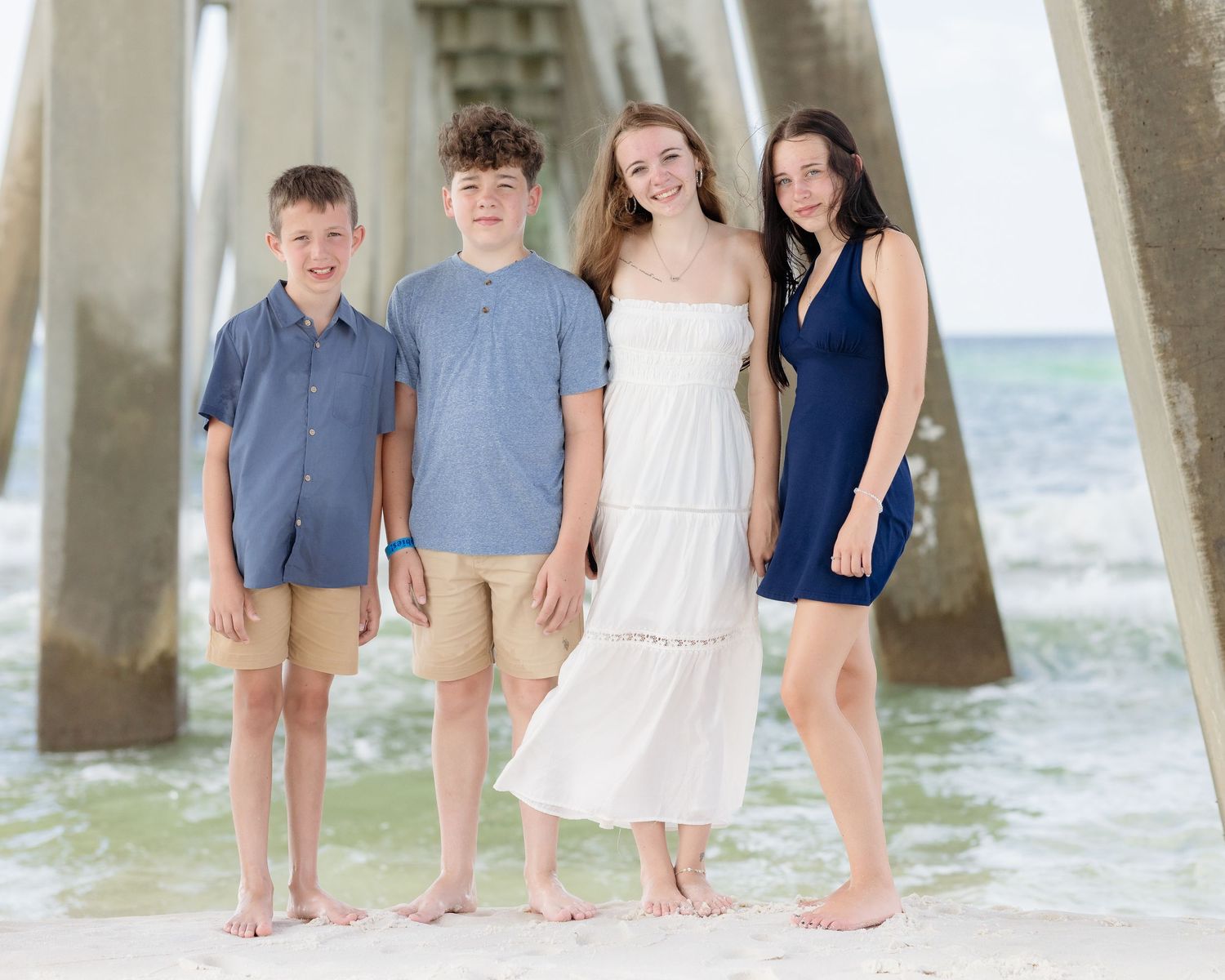 Four siblings pose barefoot on a sandy beach near a pier, wearing coordinated blue and white outfits on a sunny day.