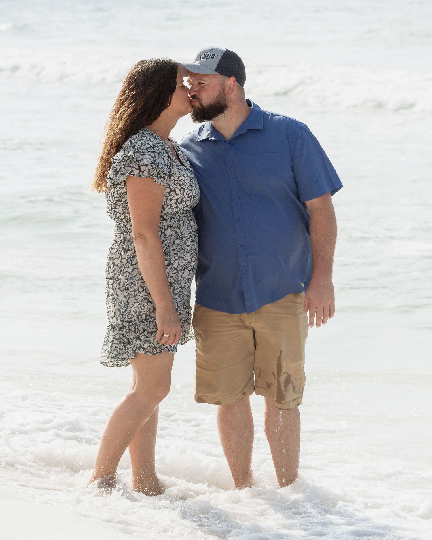 Couple sharing a romantic kiss on a sandy beach with ocean waves, woman in floral dress and man in blue shirt.