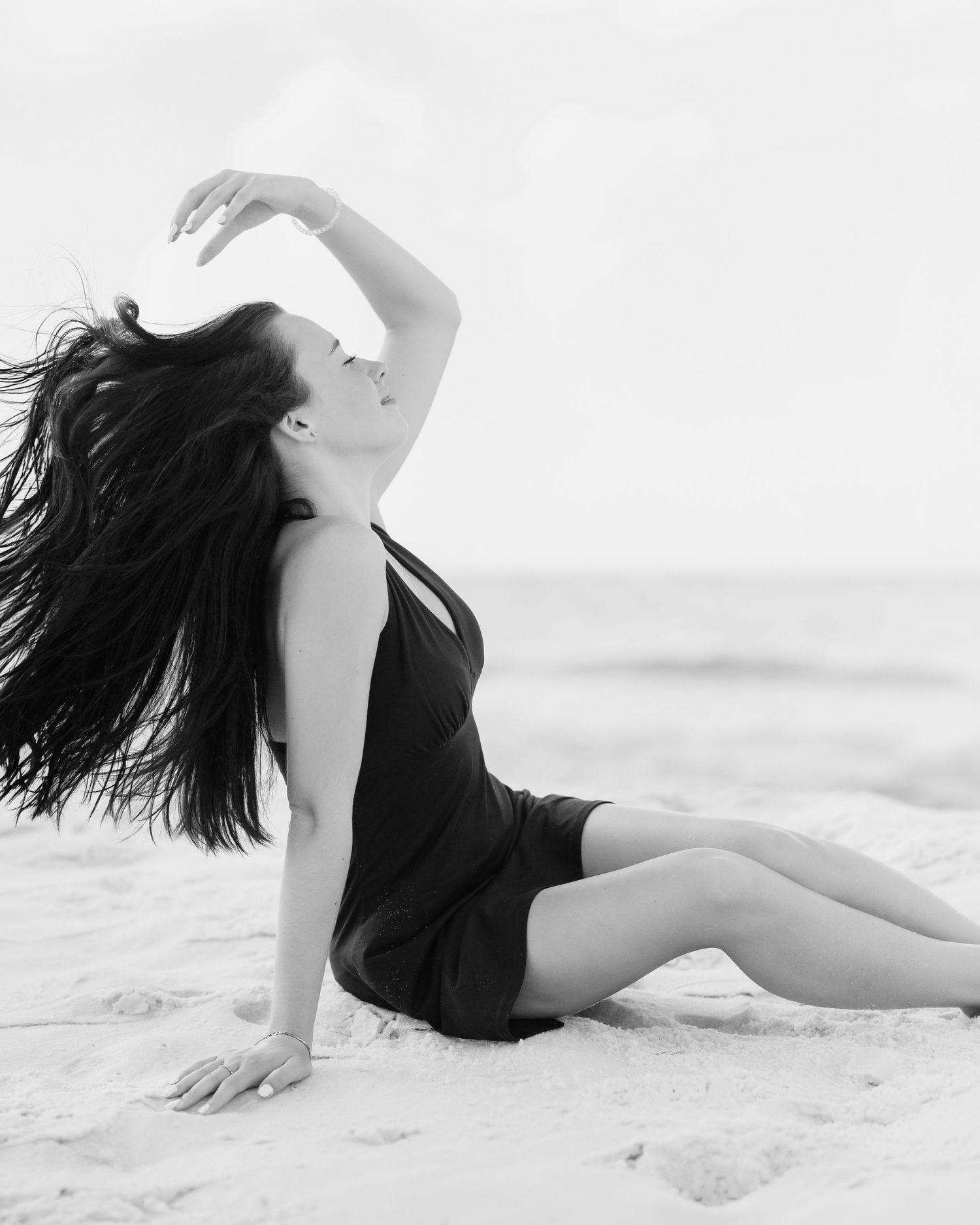 Black and white photo of a woman in a black dress sitting on a beach, hair flowing in the wind.