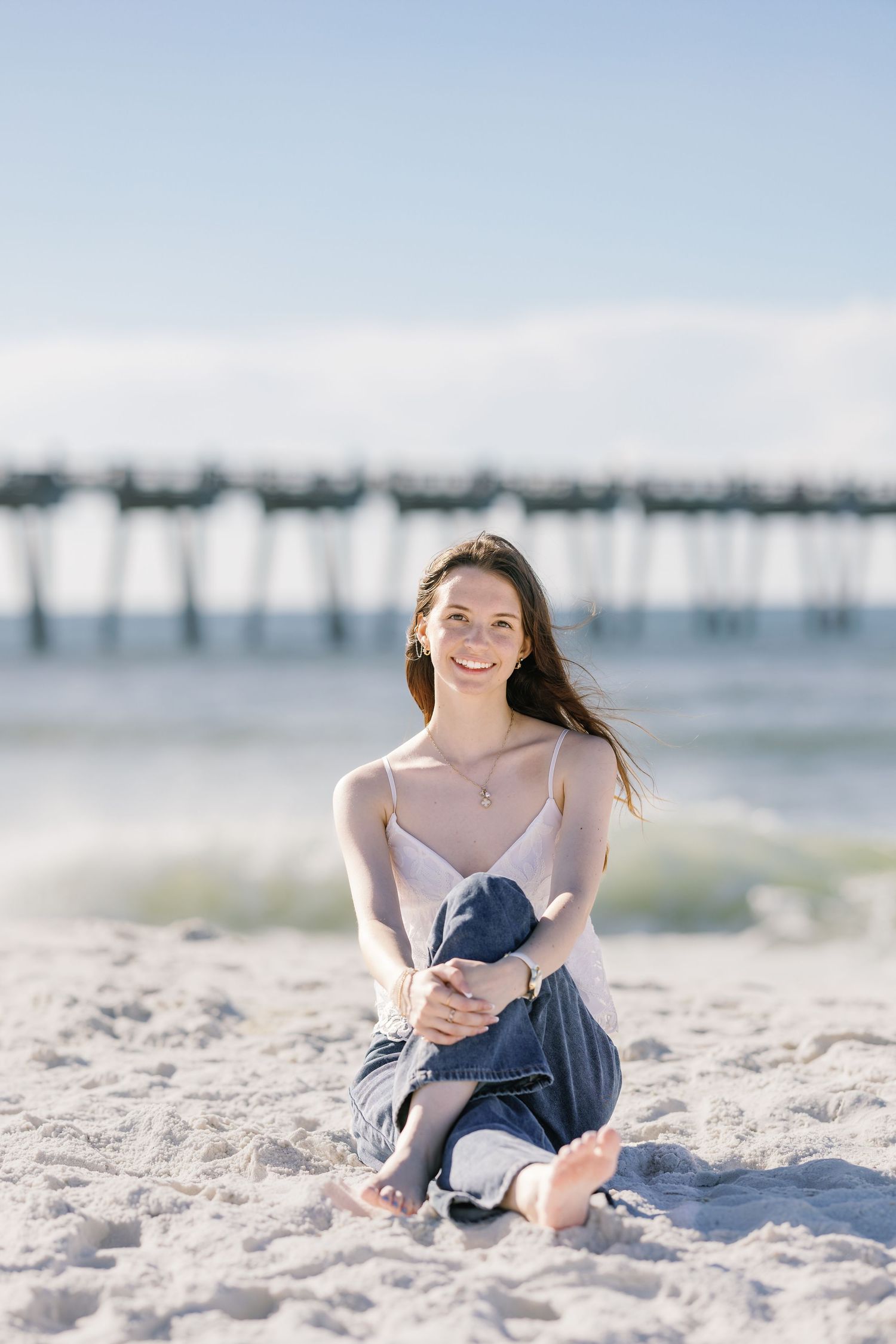 Young woman smiling on sandy beach, sitting barefoot in jeans and white top with pier in background.