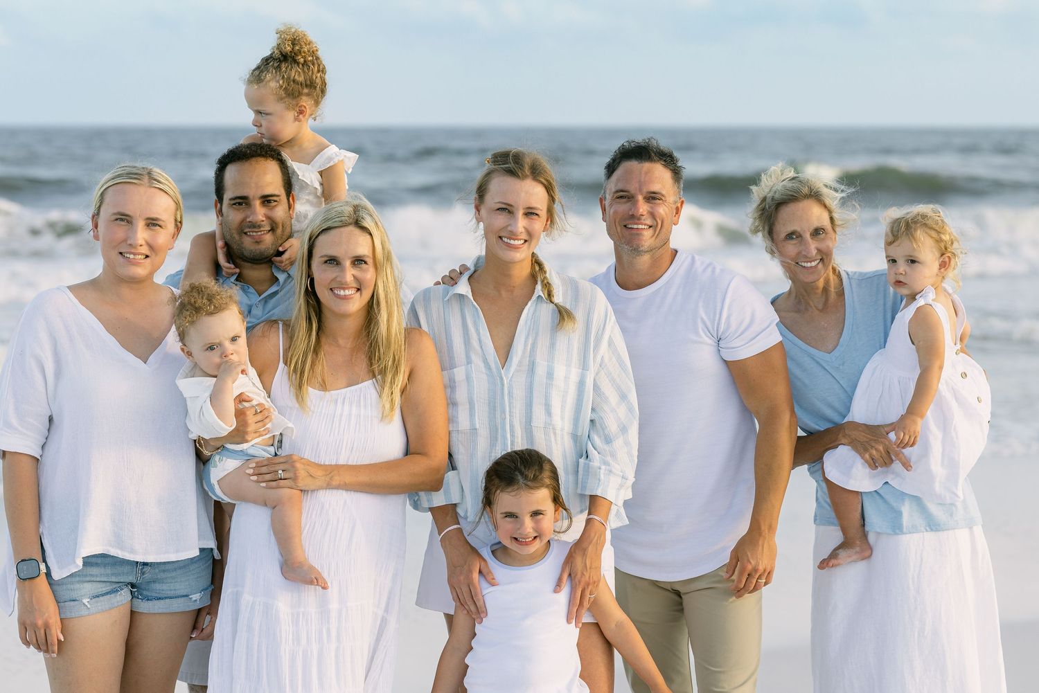 Large family group smiling together on a sunny beach, dressed in white and blue outfits, with ocean waves in the background.