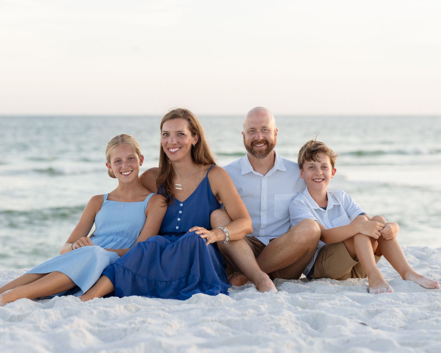 Family of four smiling together on a white sand beach with calm ocean waves in the background.
