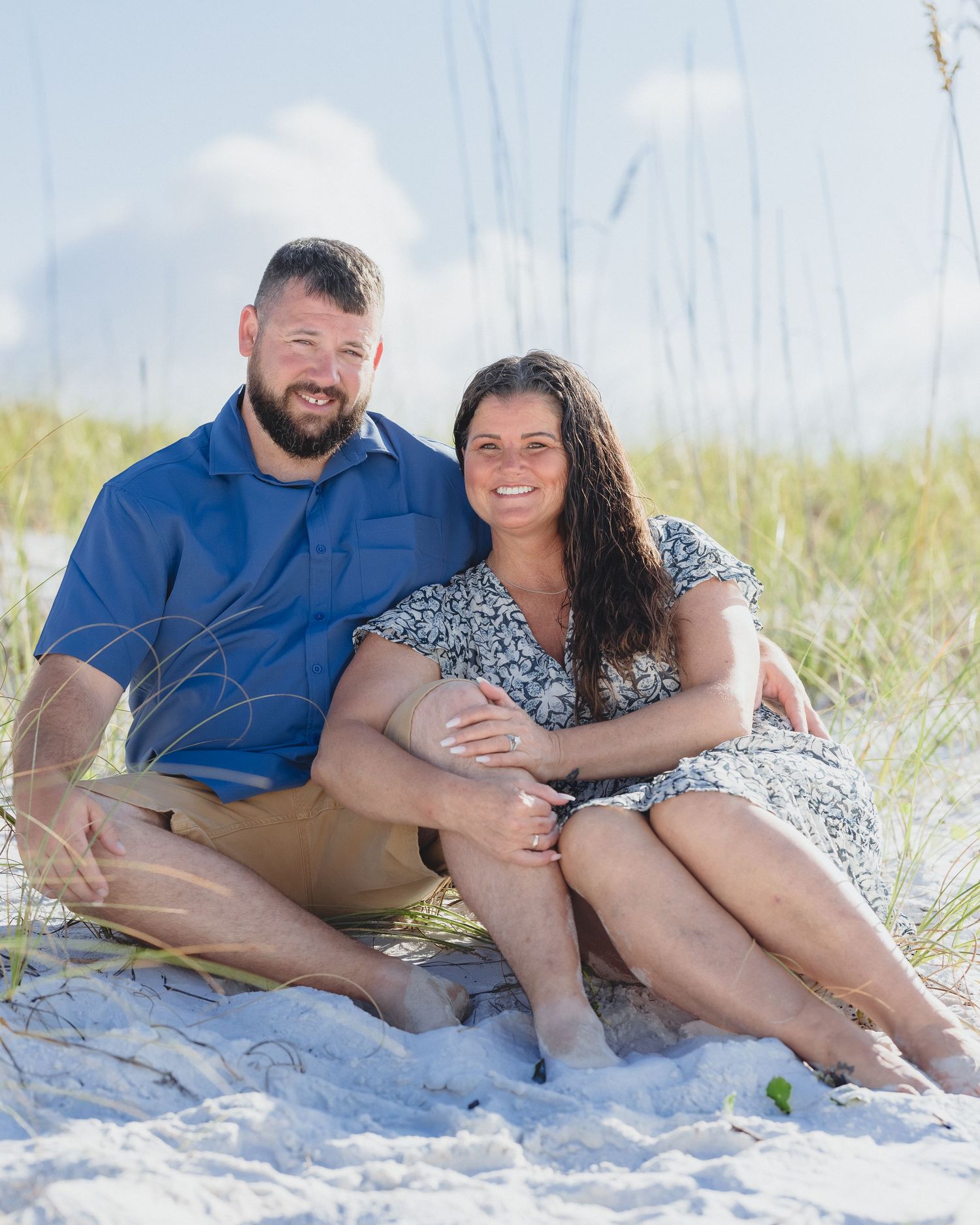 Couple smiling and sitting together on white sand beach dunes with sea oats and blue sky background.