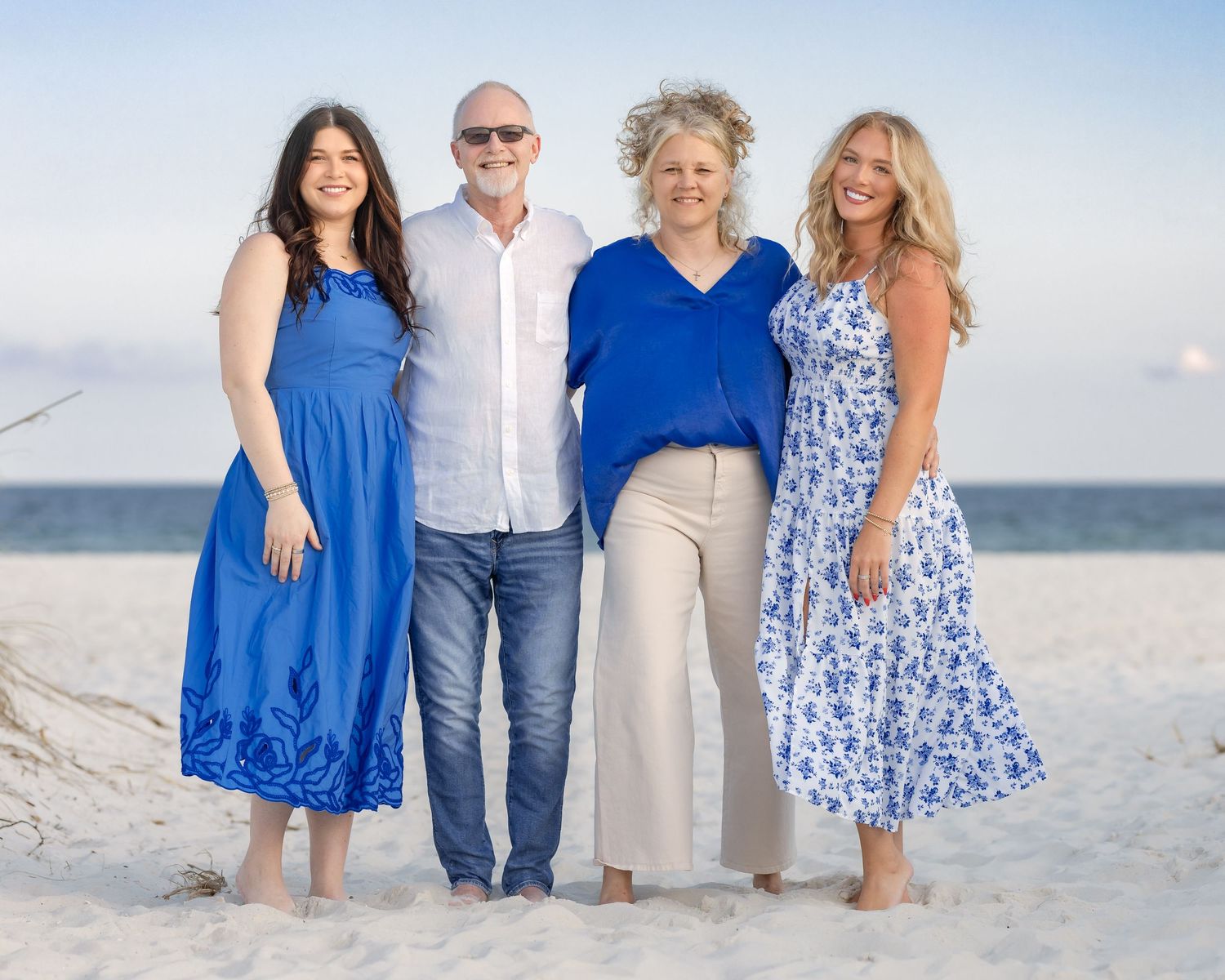 Family of four smiling together on a sandy beach at sunset, wearing coordinated blue and white outfits.