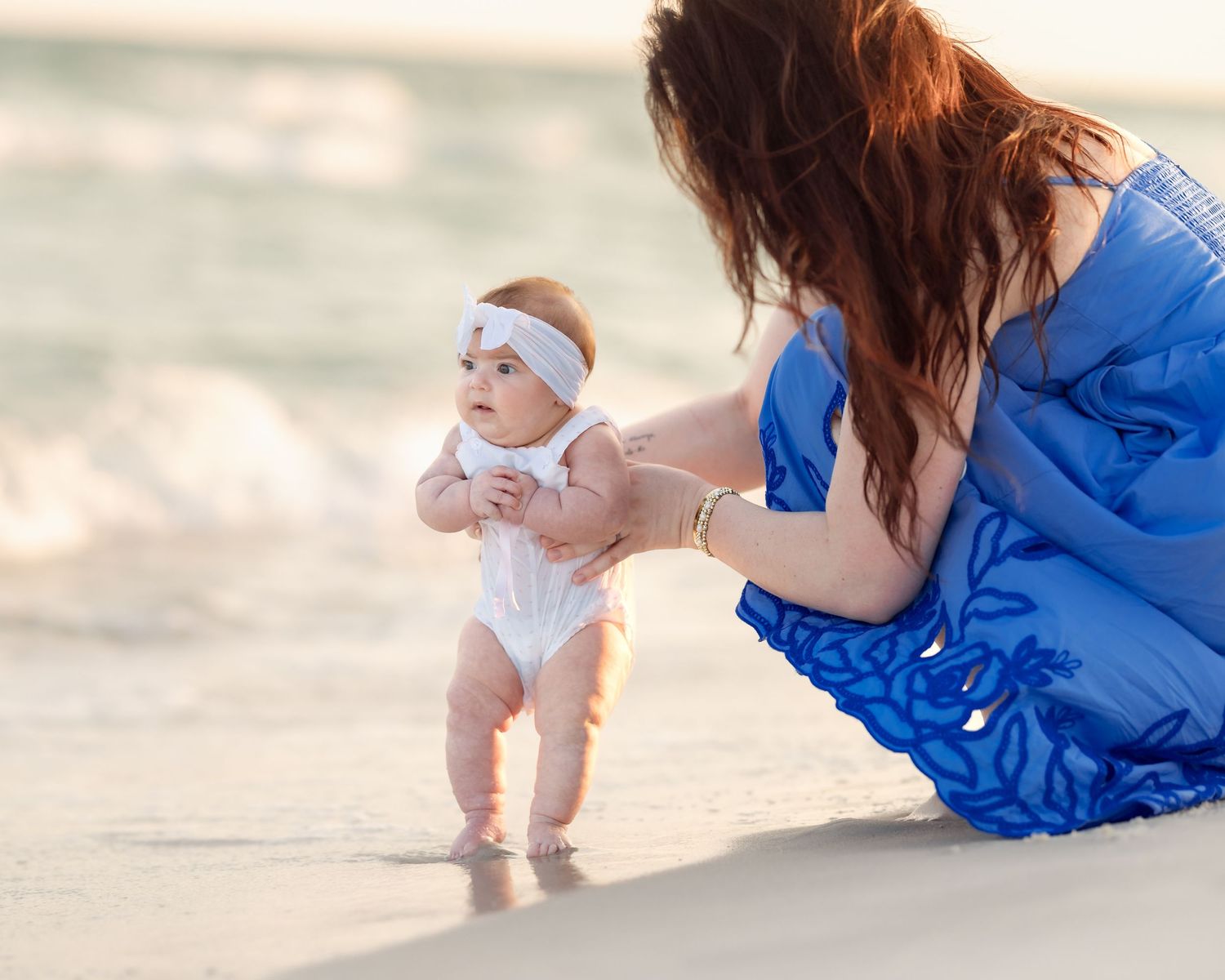 Mother in blue dress helping baby take first steps on sandy beach near ocean waves.