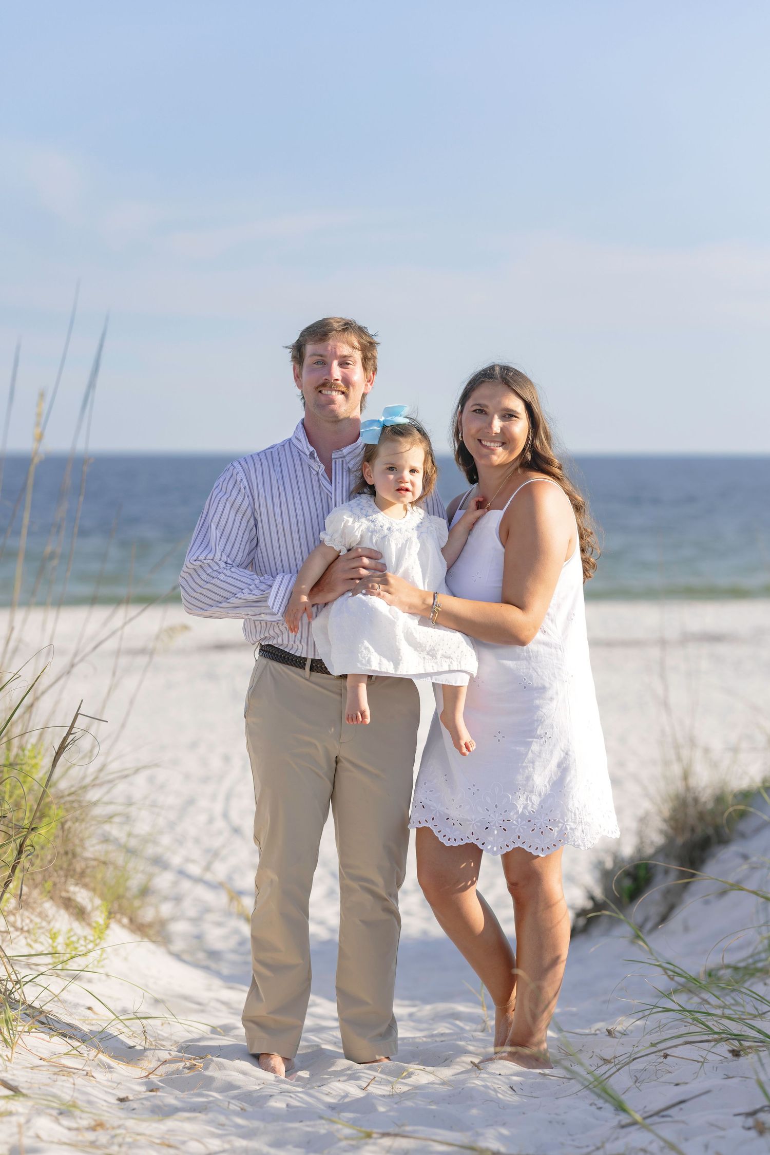 Happy family of three posing on white sand beach, parents holding toddler in white dress near sea oats.
