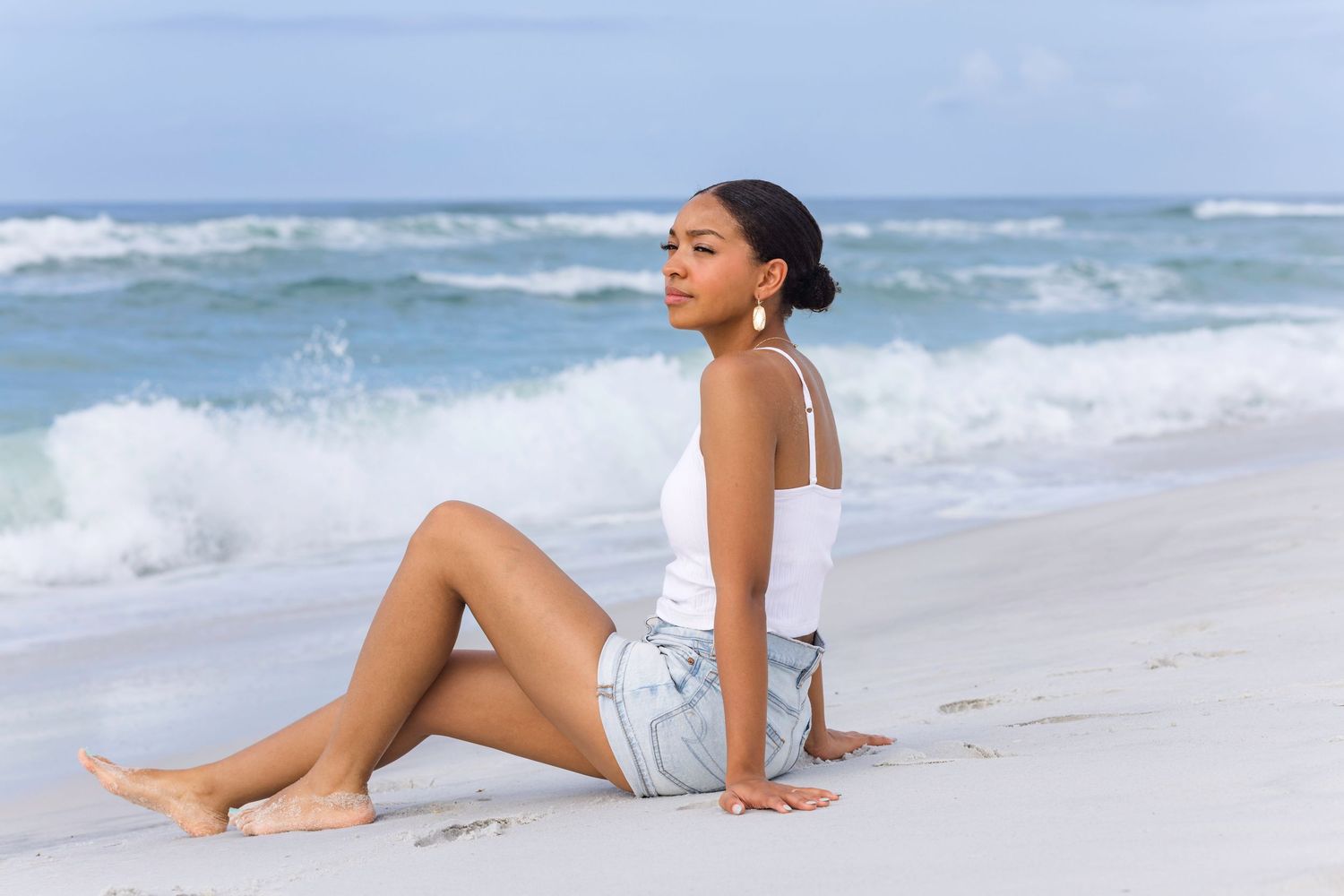 Young woman in white tank top and denim shorts sitting on a sandy beach with ocean waves behind her.