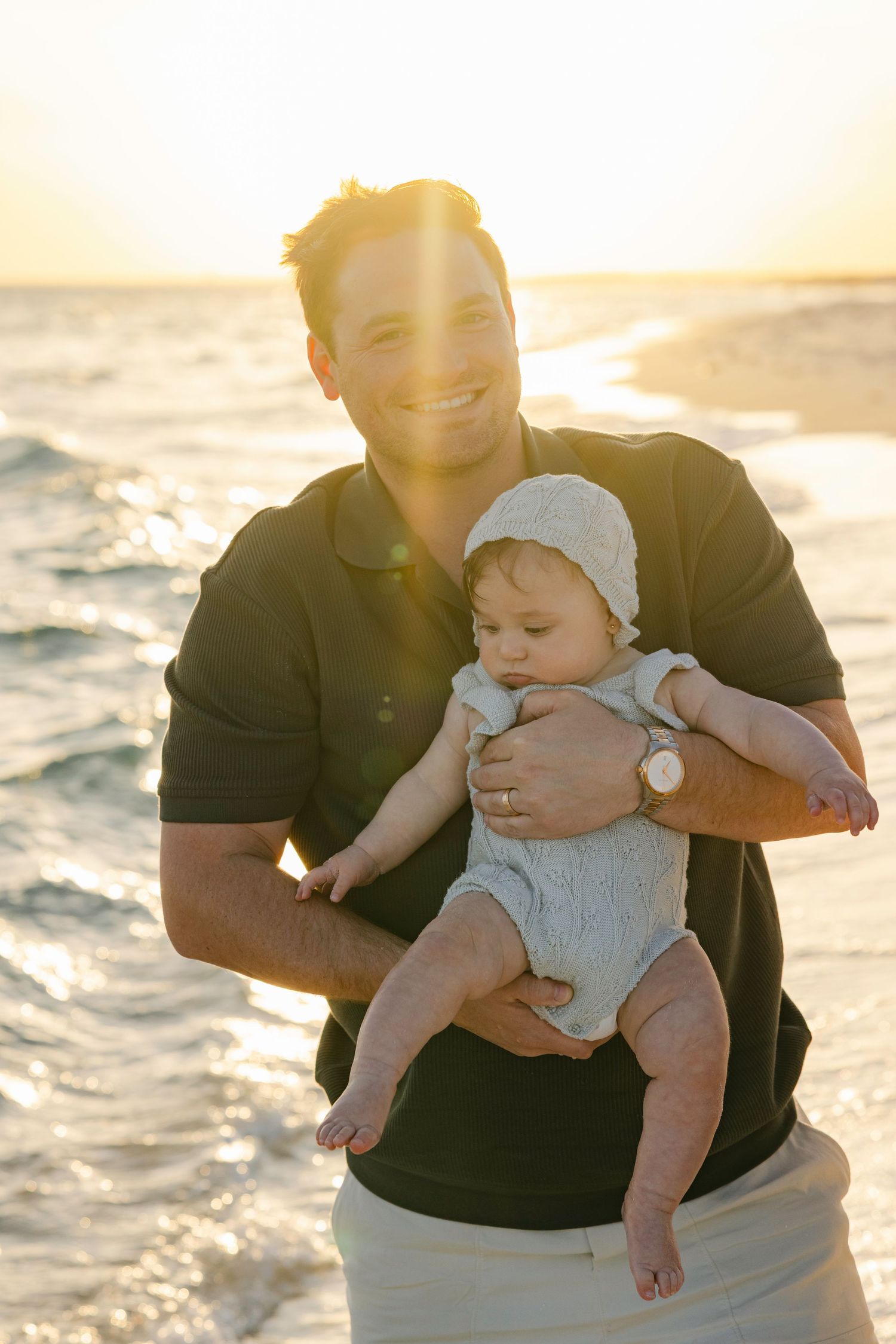 Smiling father holding baby in knit bonnet on sunny beach with ocean waves behind them.