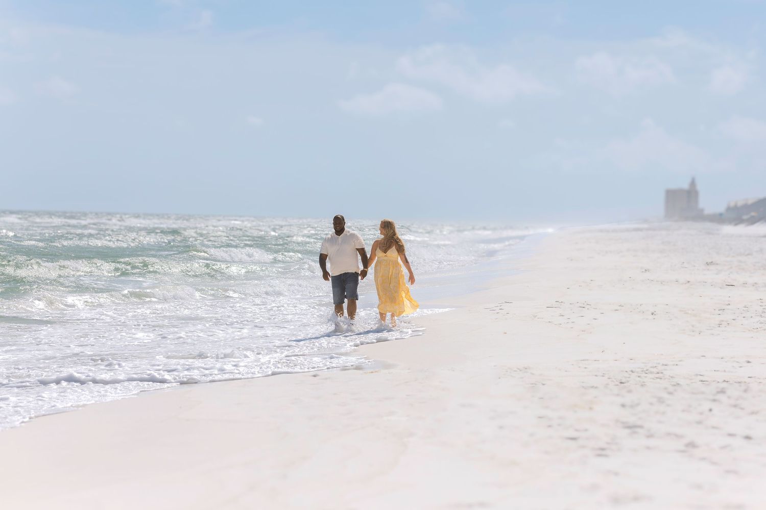 Couple walking hand in hand along a sunny beach with ocean waves and a distant lighthouse.