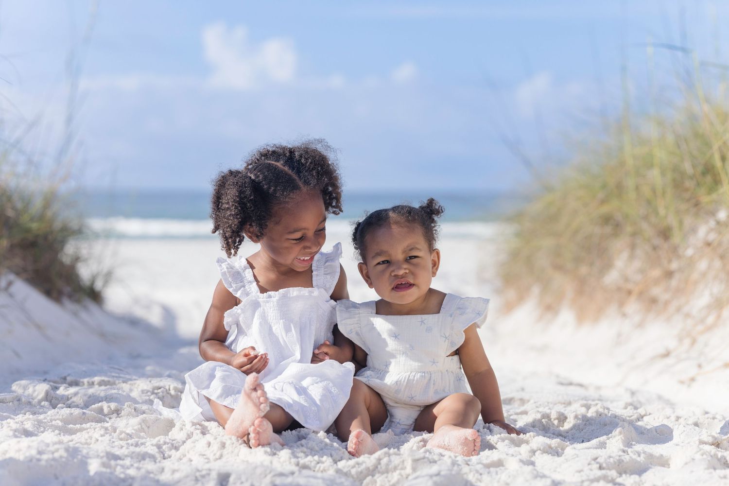 Two young girls in white dresses sitting together on white sand beach with ocean in background.