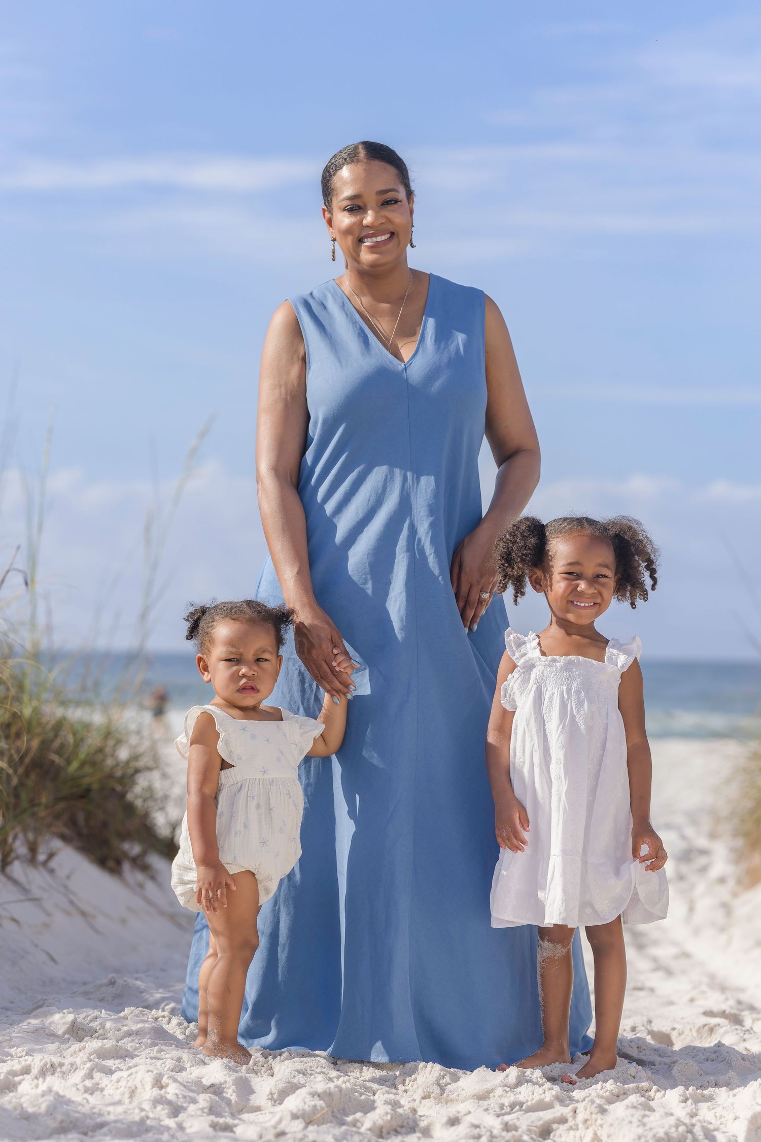 A smiling mother in a blue maxi dress poses with her two young daughters in white dresses on a sunny beach.