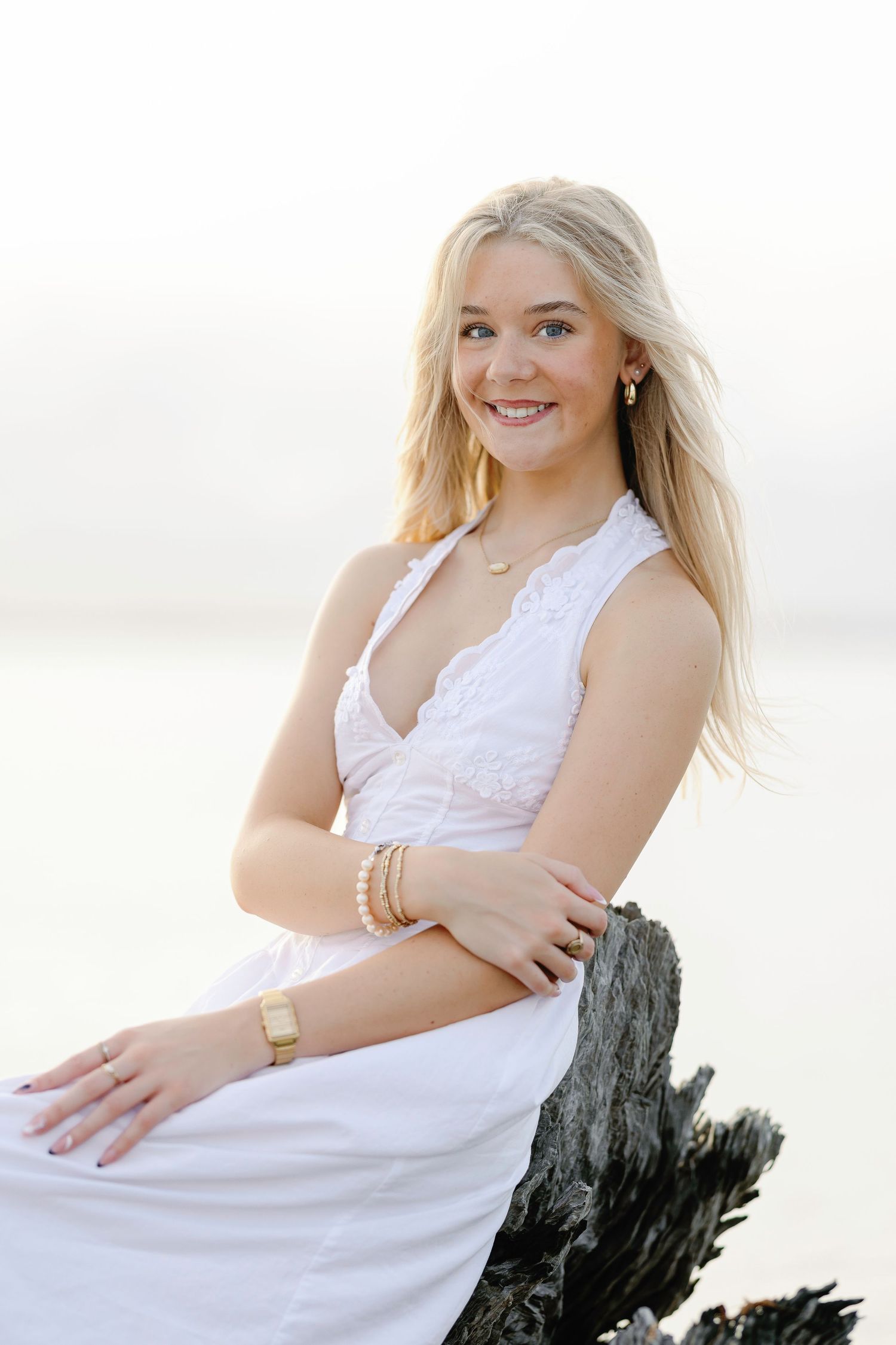Young blonde woman in white lace dress smiling at beach, wearing gold jewelry and bracelet.