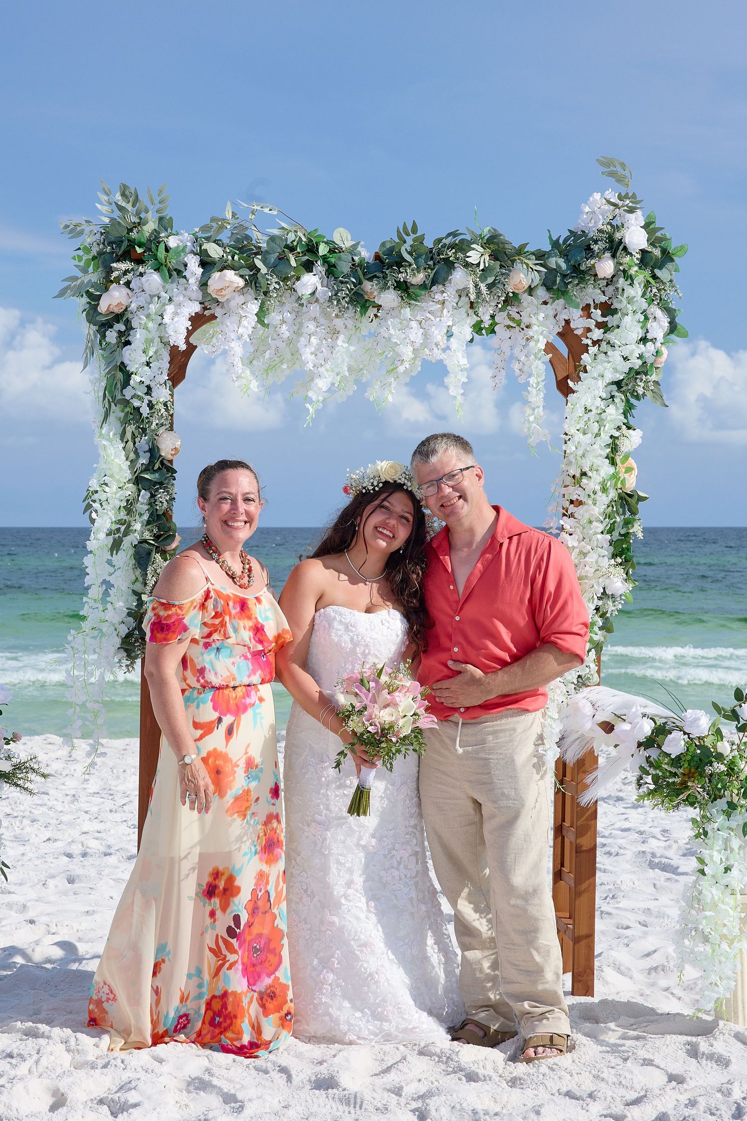Bride in white gown with bouquet poses under floral arch on beach with two guests, ocean backdrop.