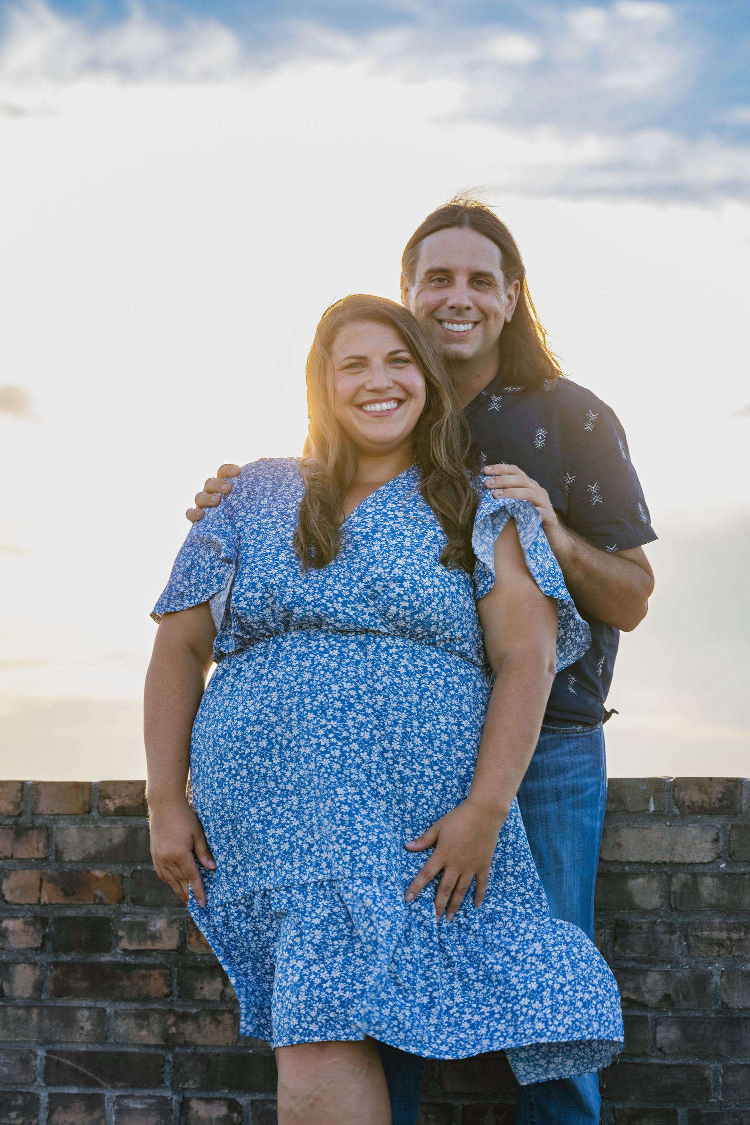 Smiling pregnant woman in blue floral dress poses with long-haired man at sunset near brick wall.