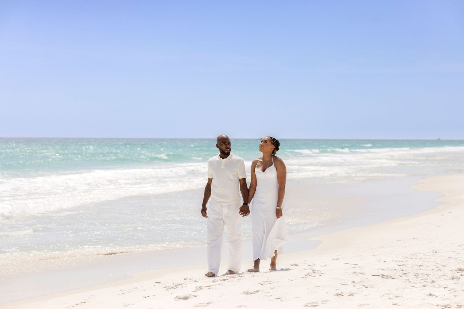 Couple in white outfits holding hands on a tropical white sand beach with turquoise ocean waters.
