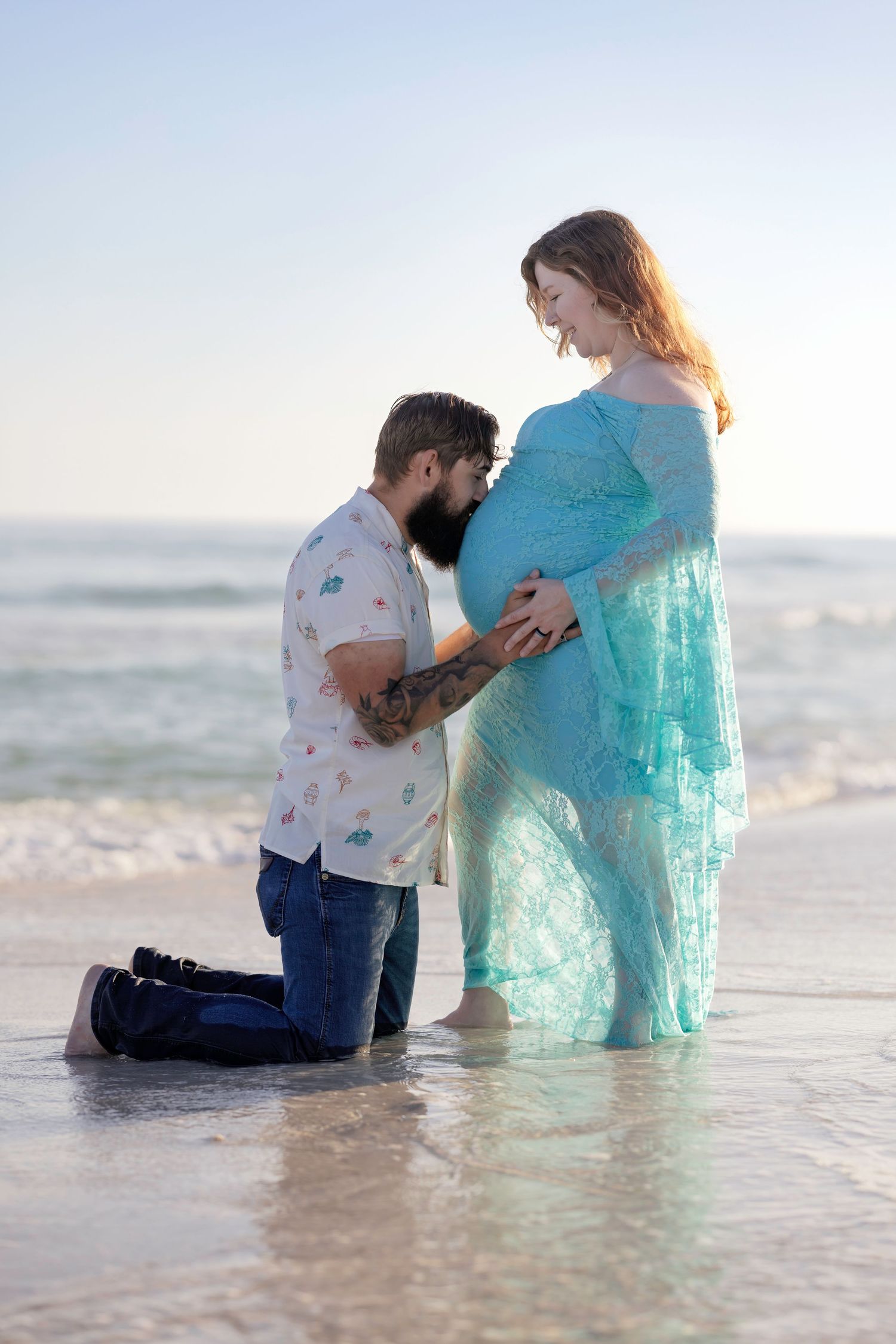 Couple sharing a tender maternity beach moment, man kneeling kissing pregnant womans belly in teal lace gown at sunset.