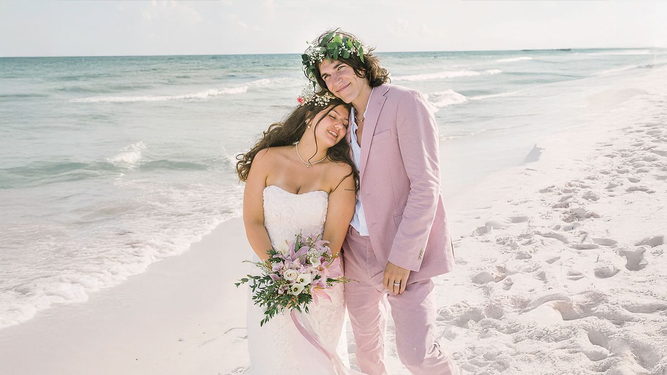 Couple in wedding attire embracing on a white sand beach, groom in pink suit, bride in white gown holding floral bouquet.