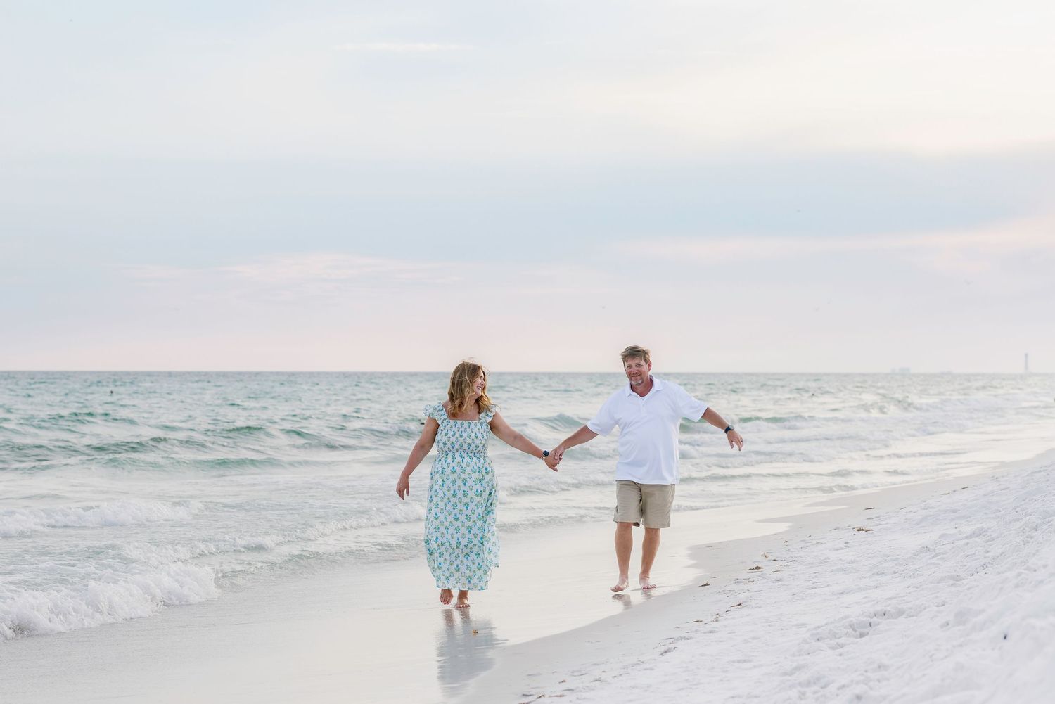 Couple holding hands walking along a white sandy beach with gentle ocean waves and a pastel sky.