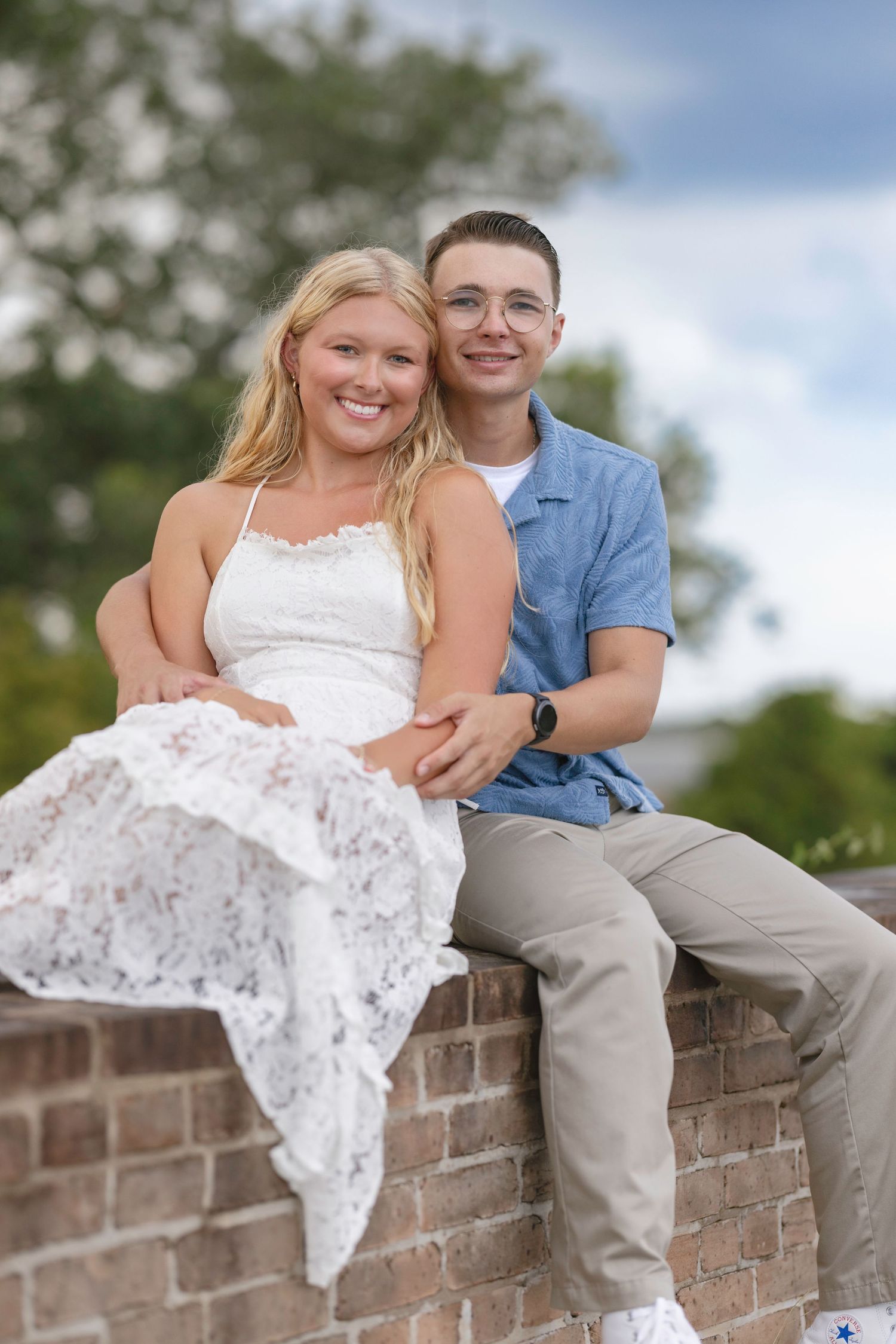 A smiling young couple sitting together on a brick wall outdoors, woman in white lace dress, man in blue shirt.