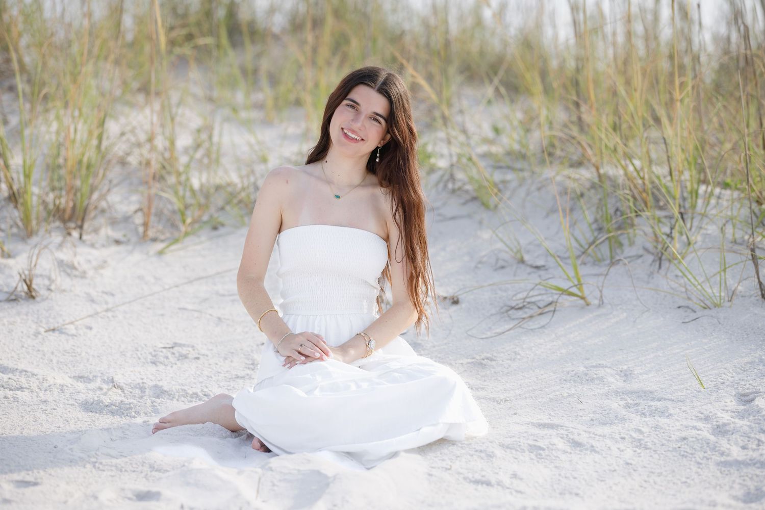 A smiling young woman in a white strapless dress sits relaxed on sandy beach dunes surrounded by grass.