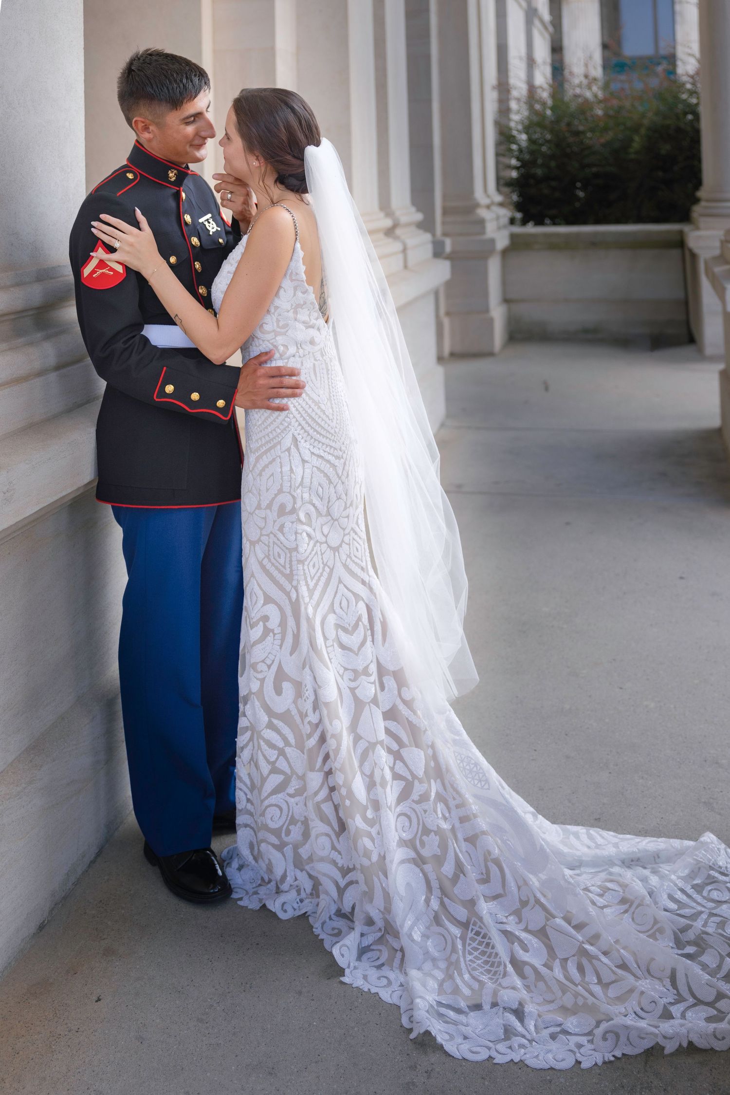 Marine groom in dress blues embraces bride in lace gown with long veil against stone columns.