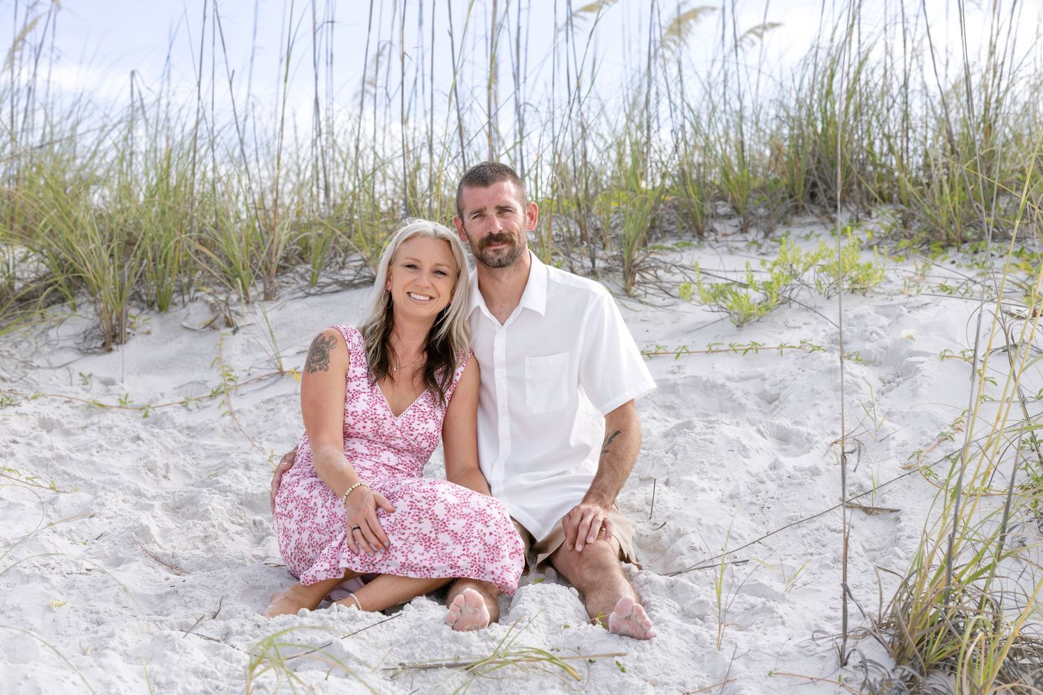 Couple smiling together on white sand beach dunes with sea oats, woman in pink floral dress, man in white shirt.